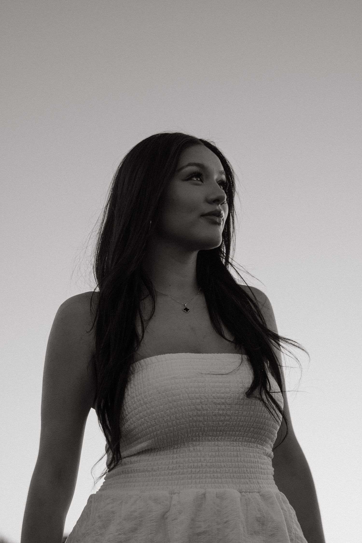 Black and white portrait of a young woman in a strapless dress against a clear sky.