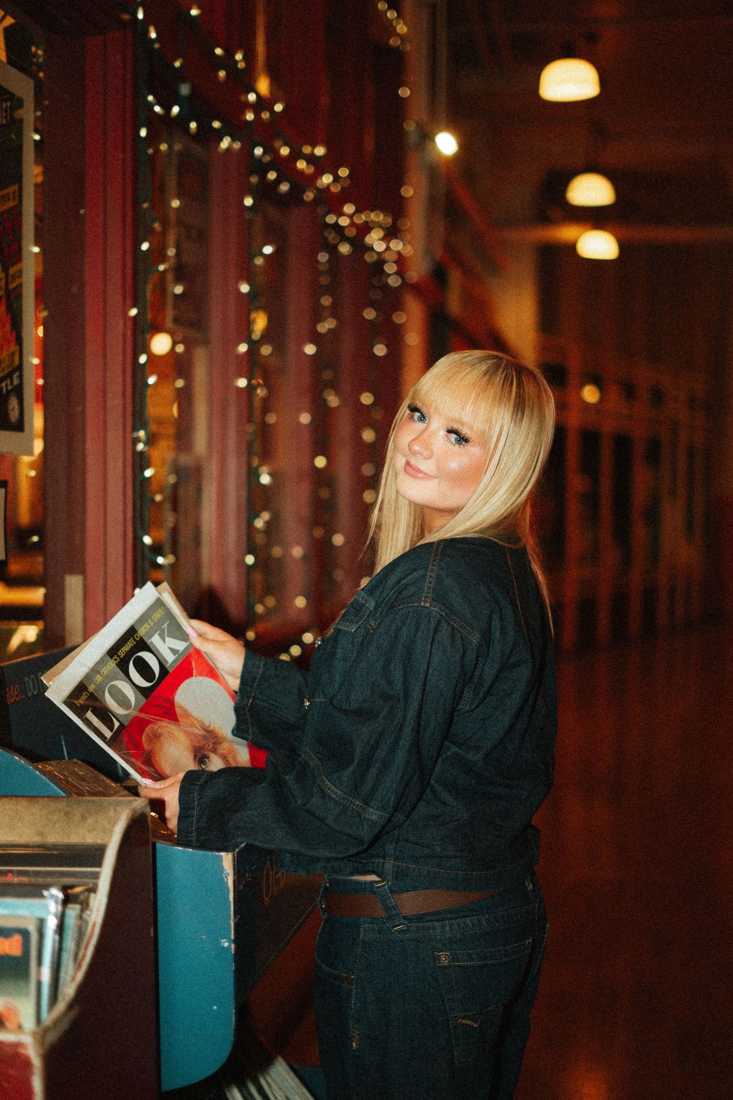 Smiling blonde woman in denim jacket browsing vinyl records at a jukebox in a warmly lit venue.