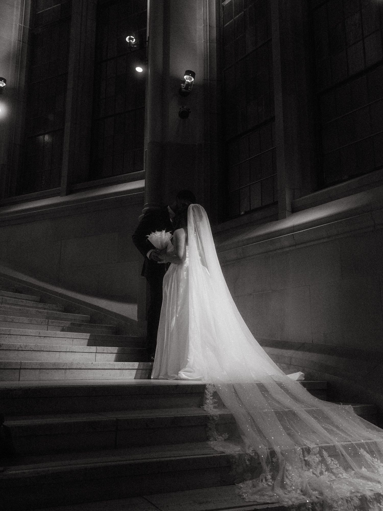 A bride and groom kiss on grand stone stairs in a dramatic black and white wedding portrait.
