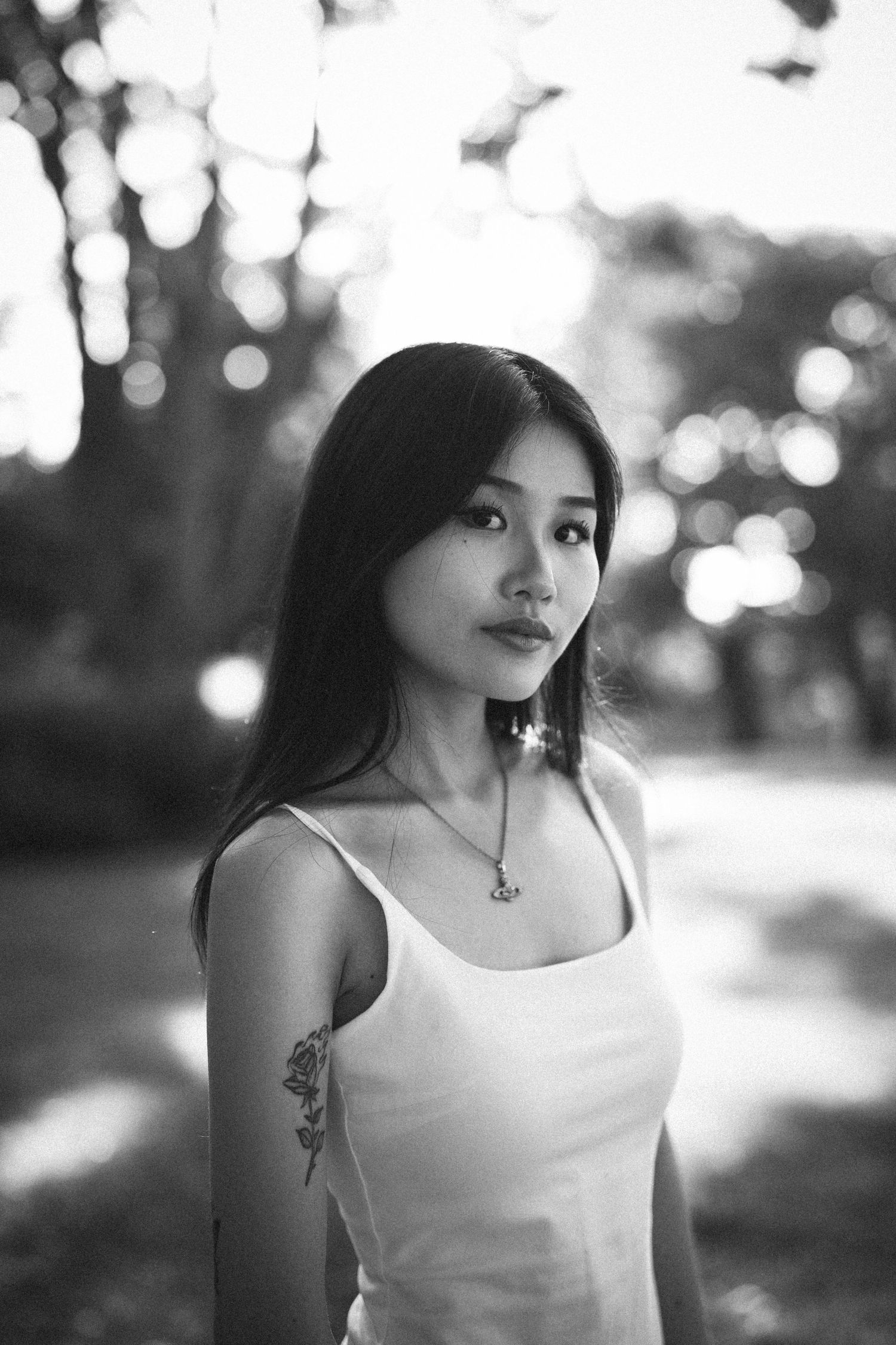 Black and white portrait of a young Asian woman with long hair, floral arm tattoo, and necklace, standing outdoors.