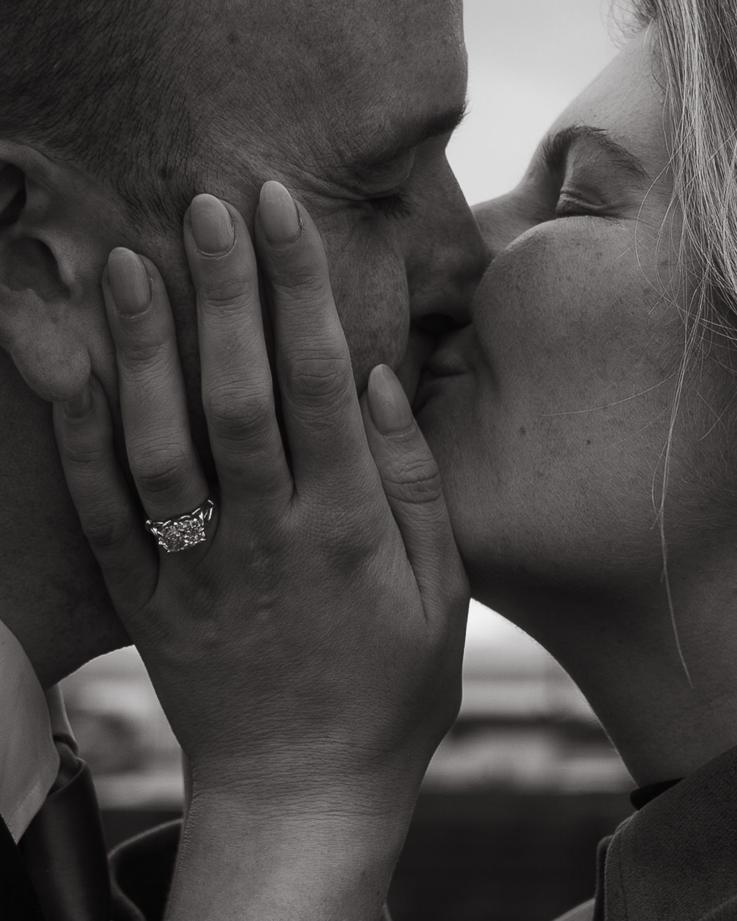 Black and white close-up of newly engaged couple kissing with diamond ring visible.