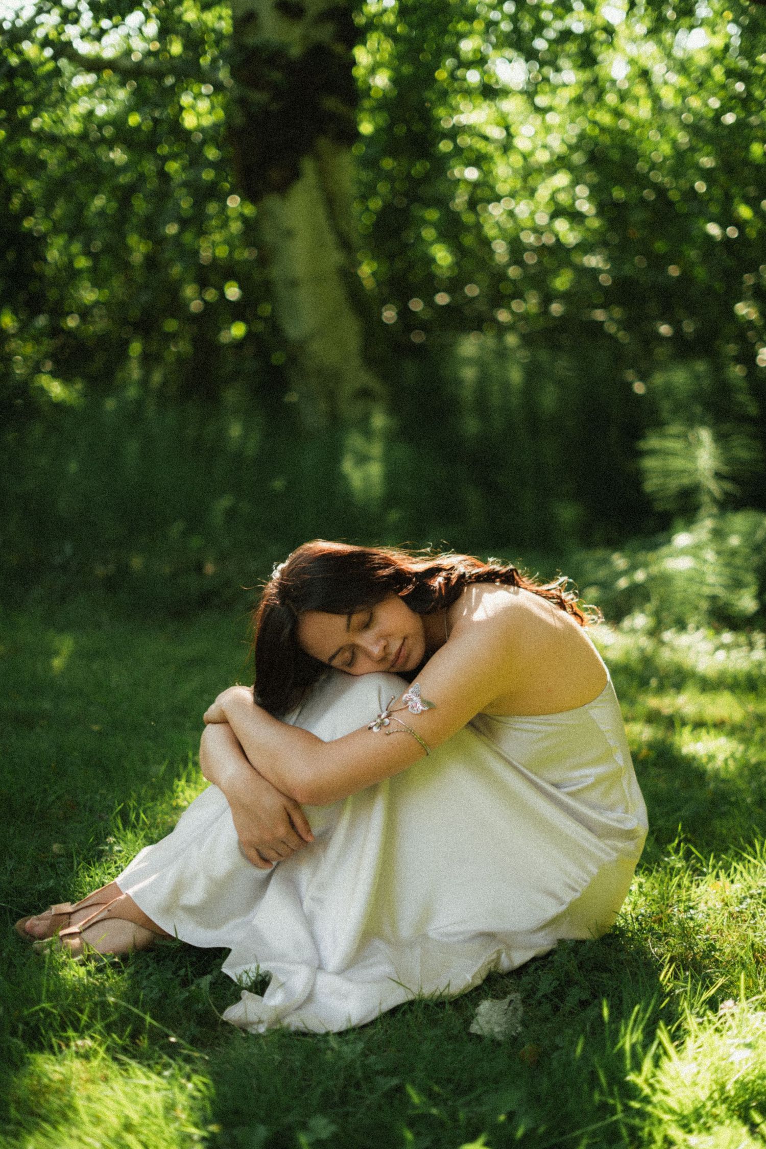 Woman in white dress sitting on grass, hugging knees, surrounded by lush green trees in soft sunlight.