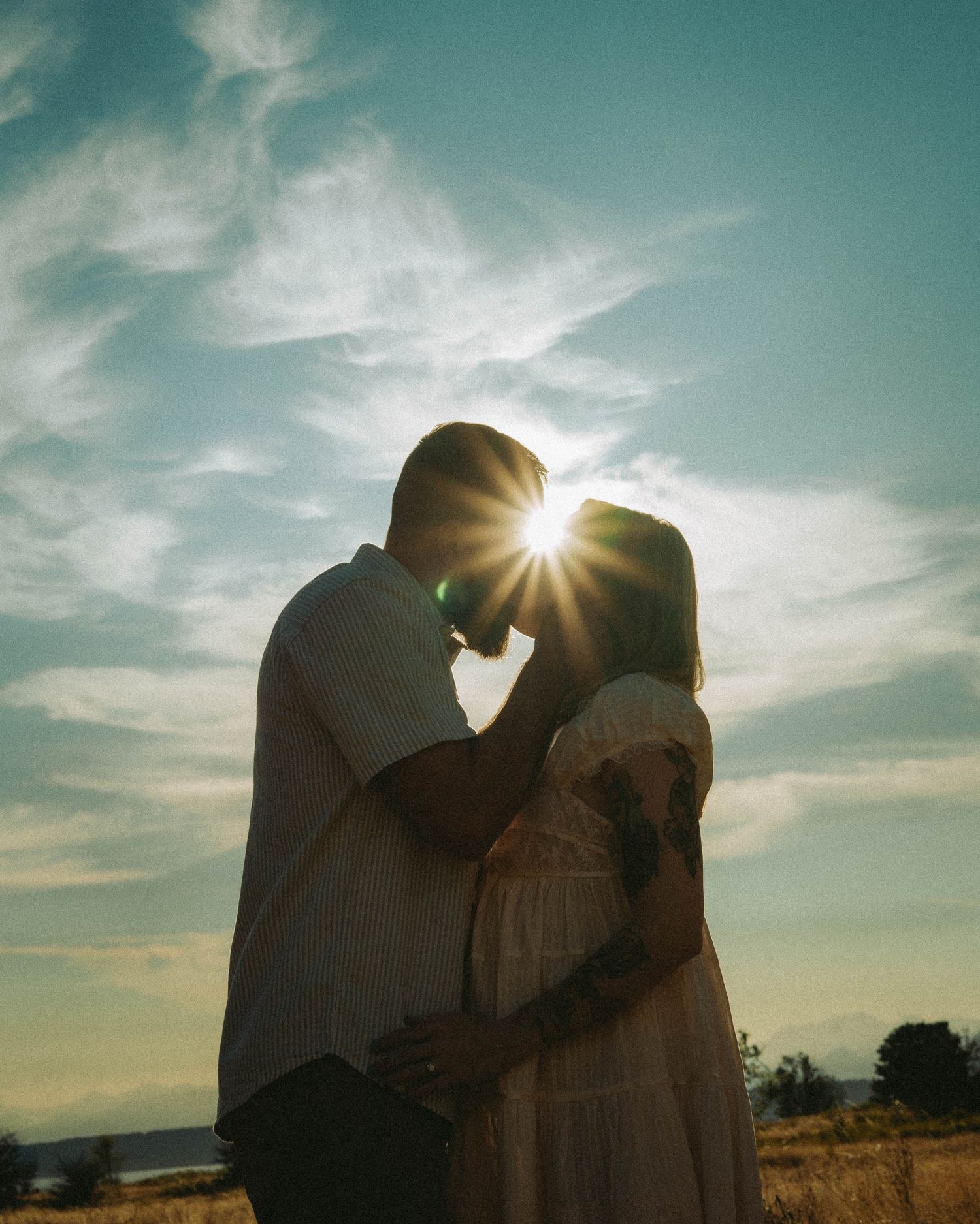 A couple shares a romantic moment silhouetted against a golden sunset, with sun rays glowing between them in an open field.