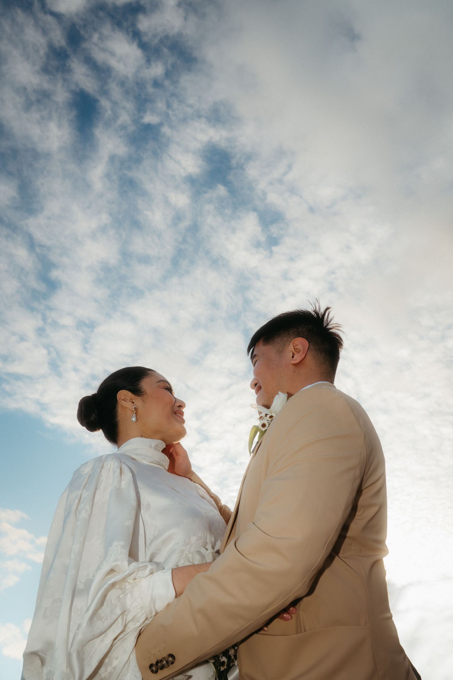 Bride and groom in formal attire gazing at each other under a blue cloudy sky.
