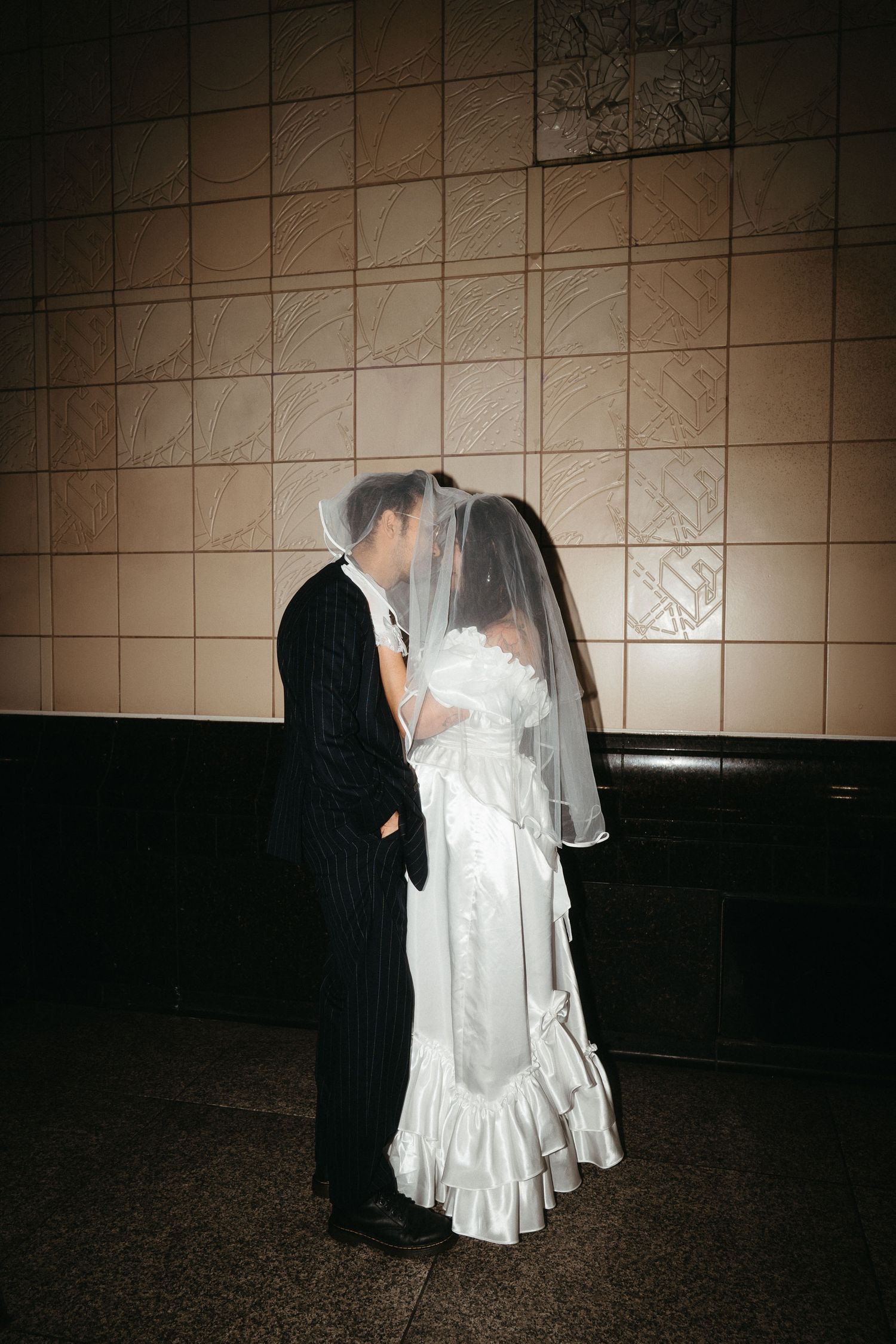 Bride and groom sharing a kiss under a veil in a tiled subway station, wearing classic wedding attire.