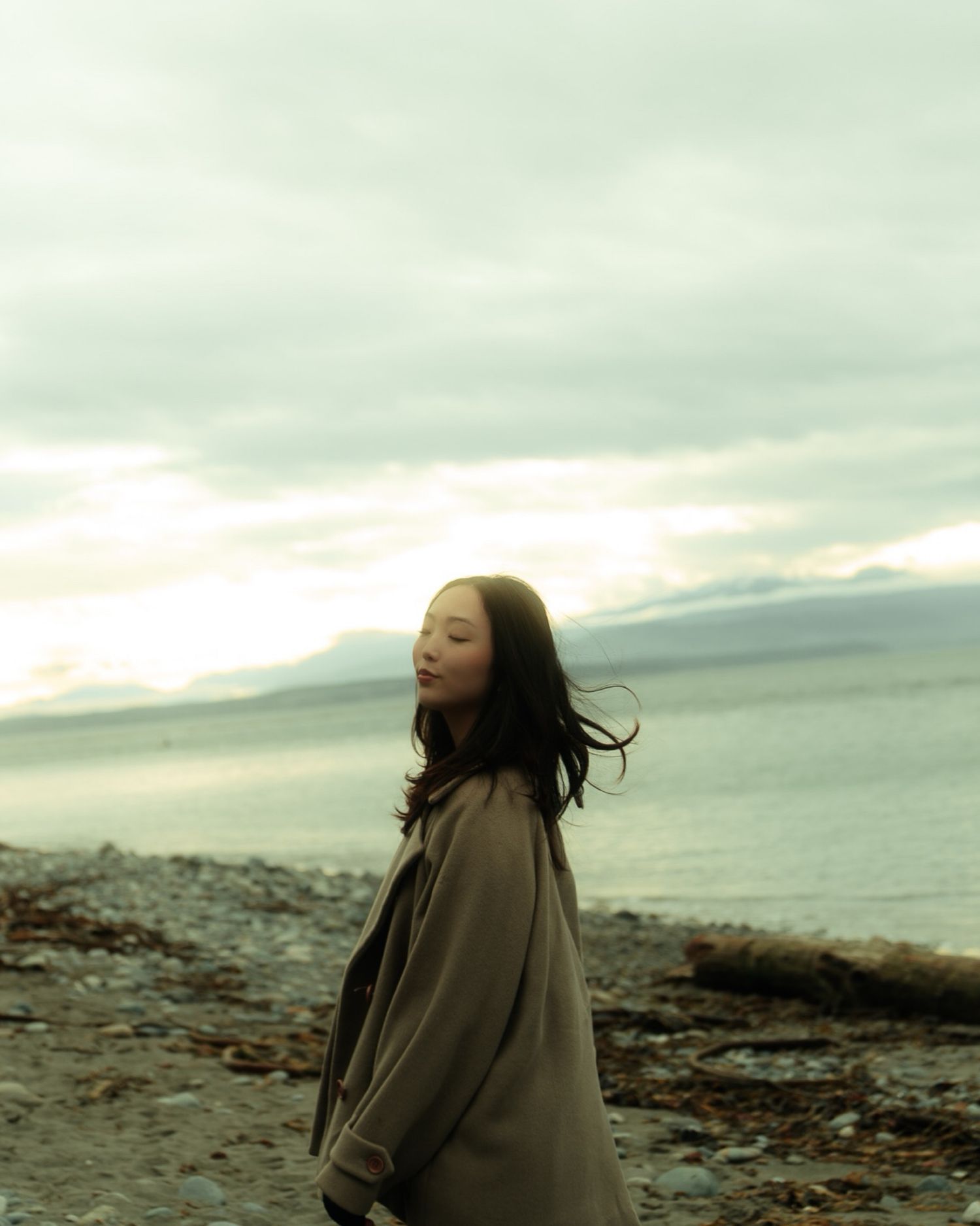 Young woman in tan coat standing on rocky beach with overcast sky and ocean.