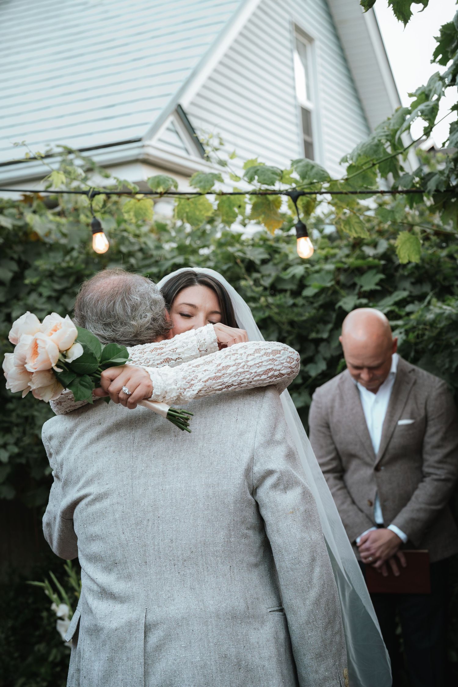 Bride in lace gown and veil hugging a guest while holding peach flowers at an outdoor wedding ceremony.
