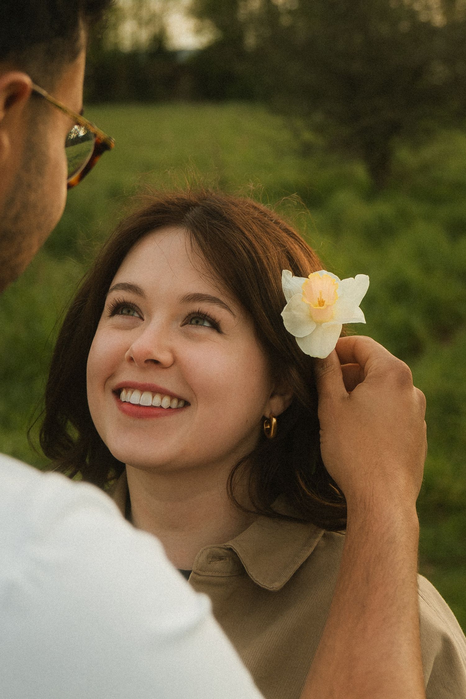 Man placing a white flower behind a smiling woman's ear in a lush green setting.