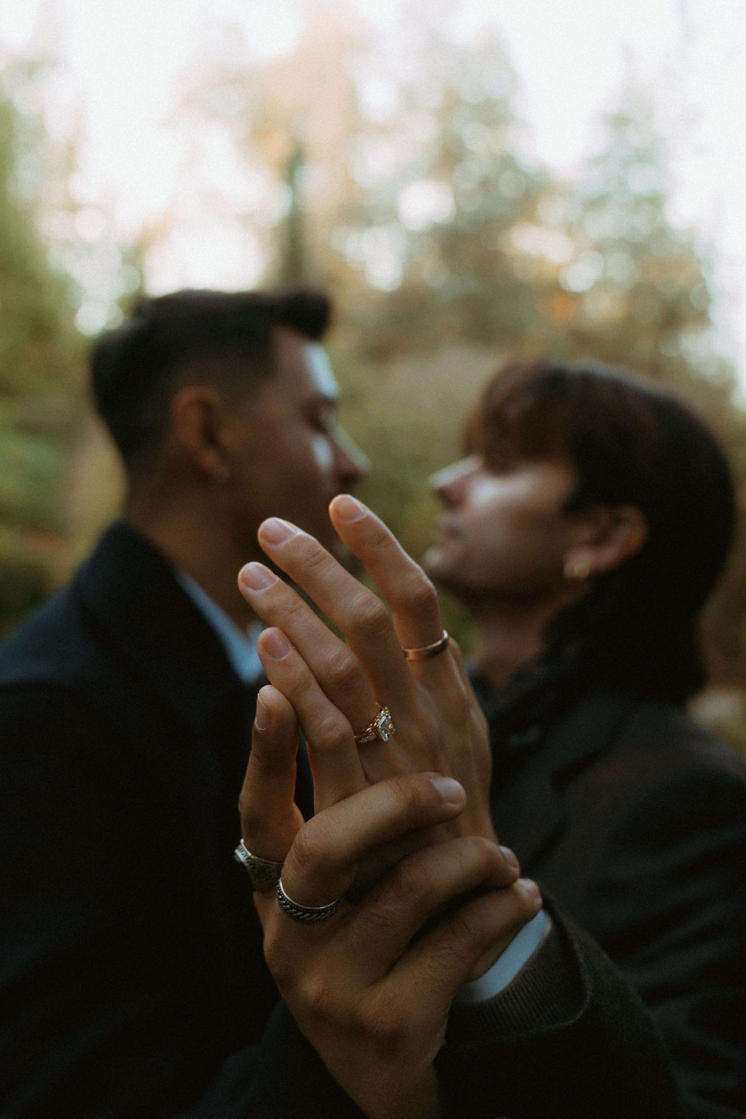 Two grooms holding hands with wedding rings in focus during outdoor forest engagement session.