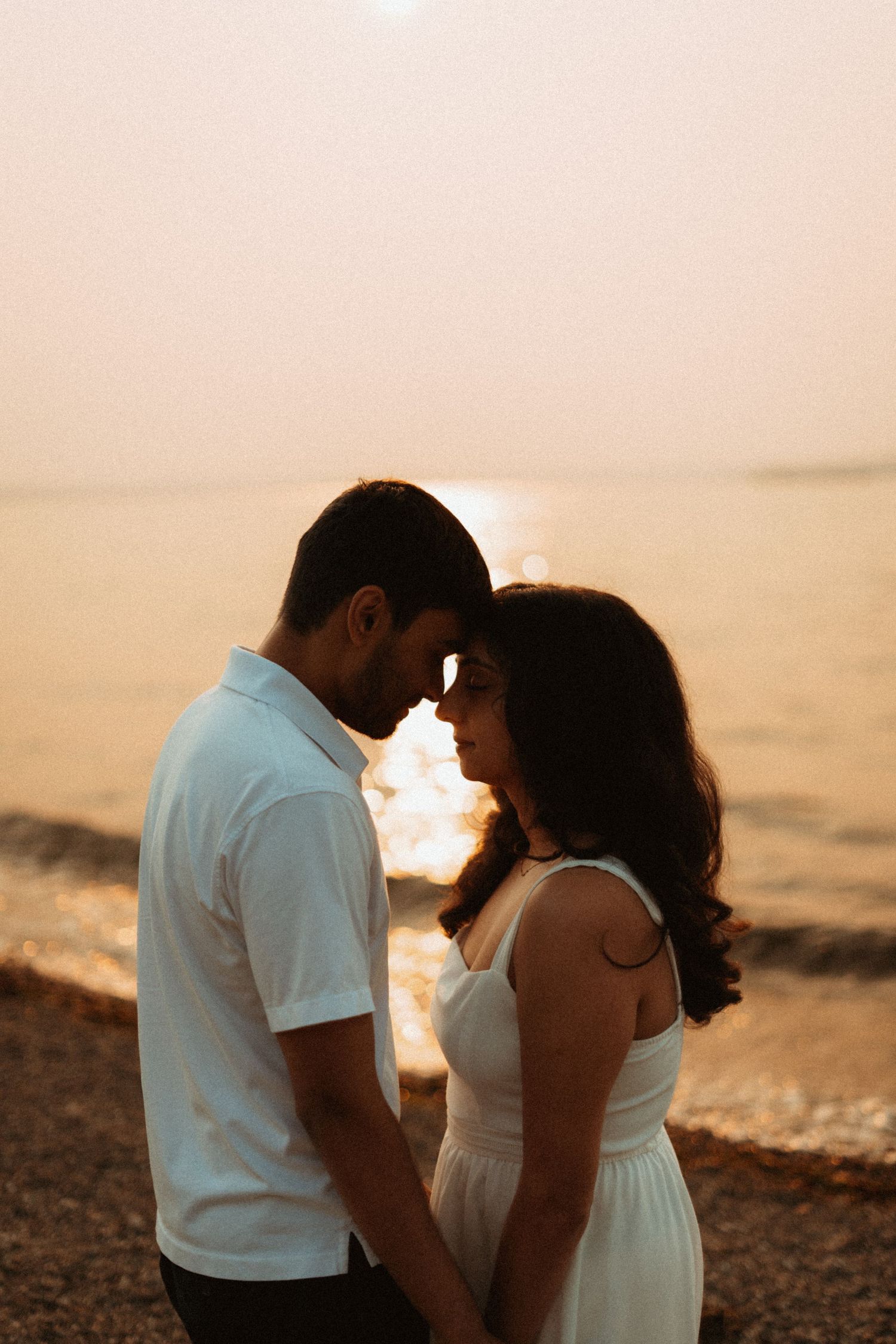 A romantic couple shares an intimate moment on a beach at sunset, with golden light reflecting off the calm ocean water.