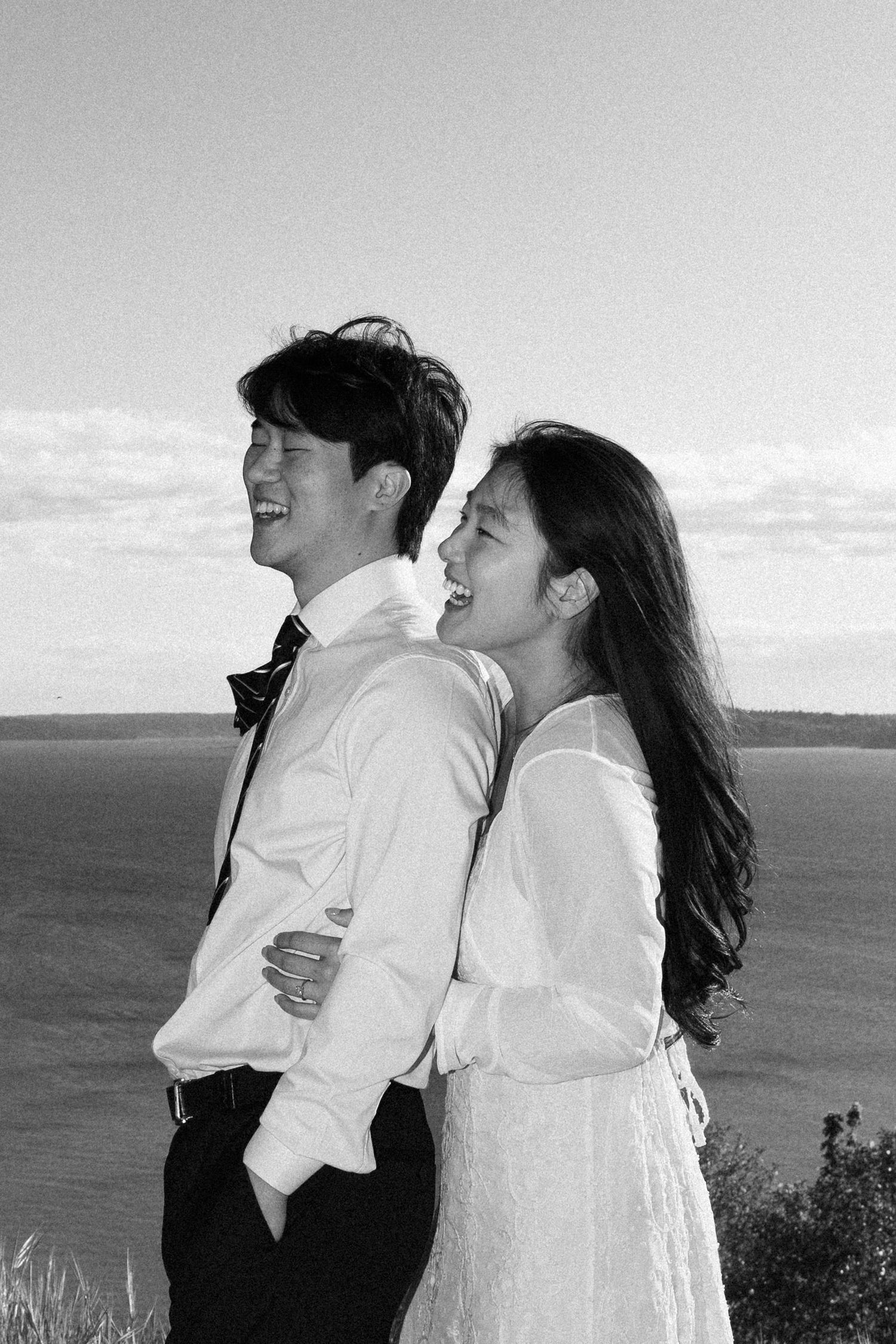 Black and white photo of a laughing couple embracing outdoors by a scenic waterfront.