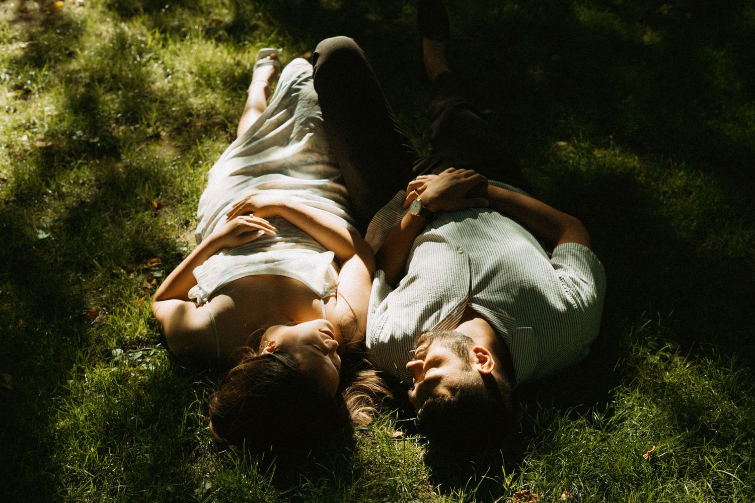 Couple lying on green grass in dappled sunlight, relaxing together outdoors in a peaceful garden setting.