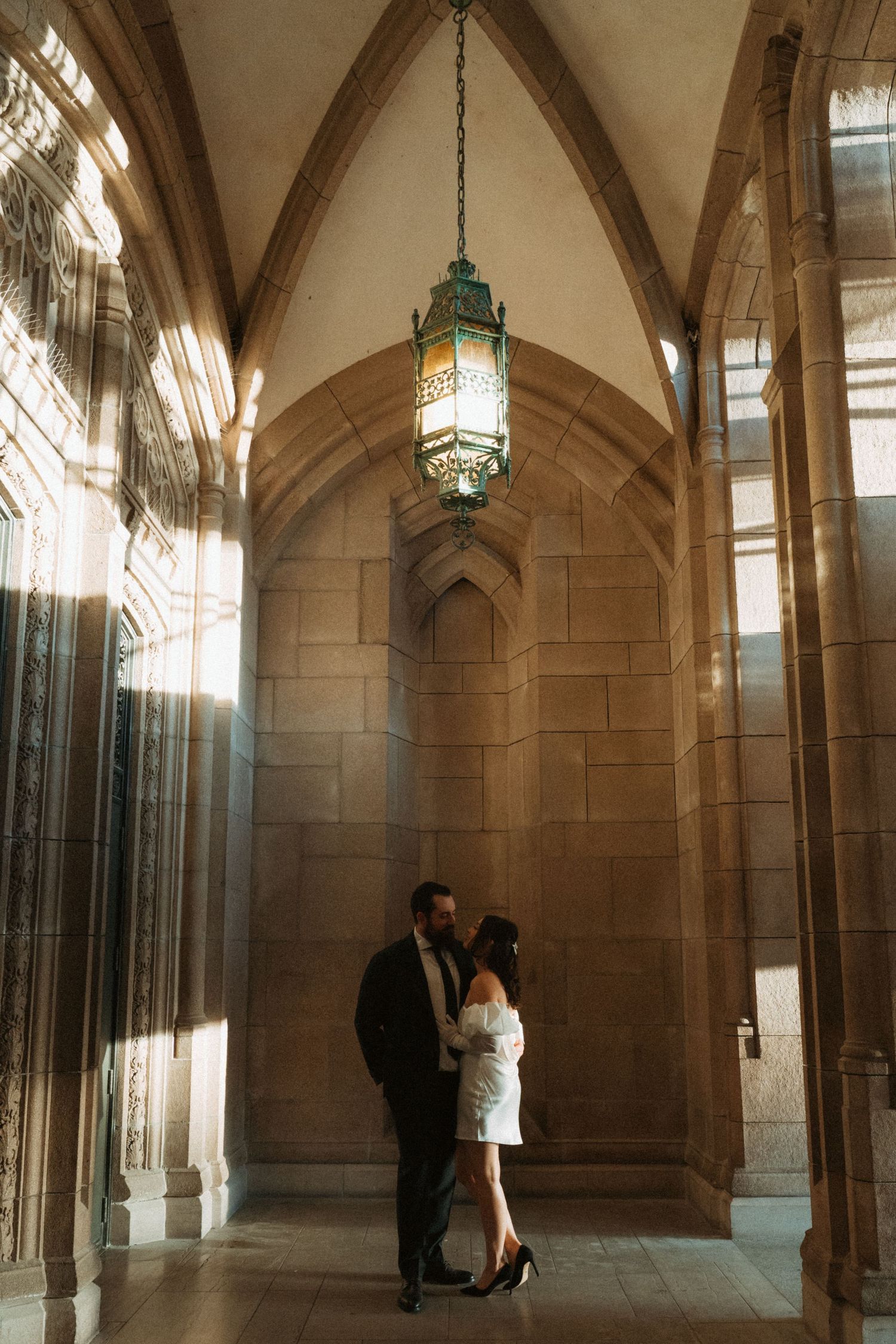 Couple embracing under gothic stone arches with a vintage lantern in a romantic indoor setting.