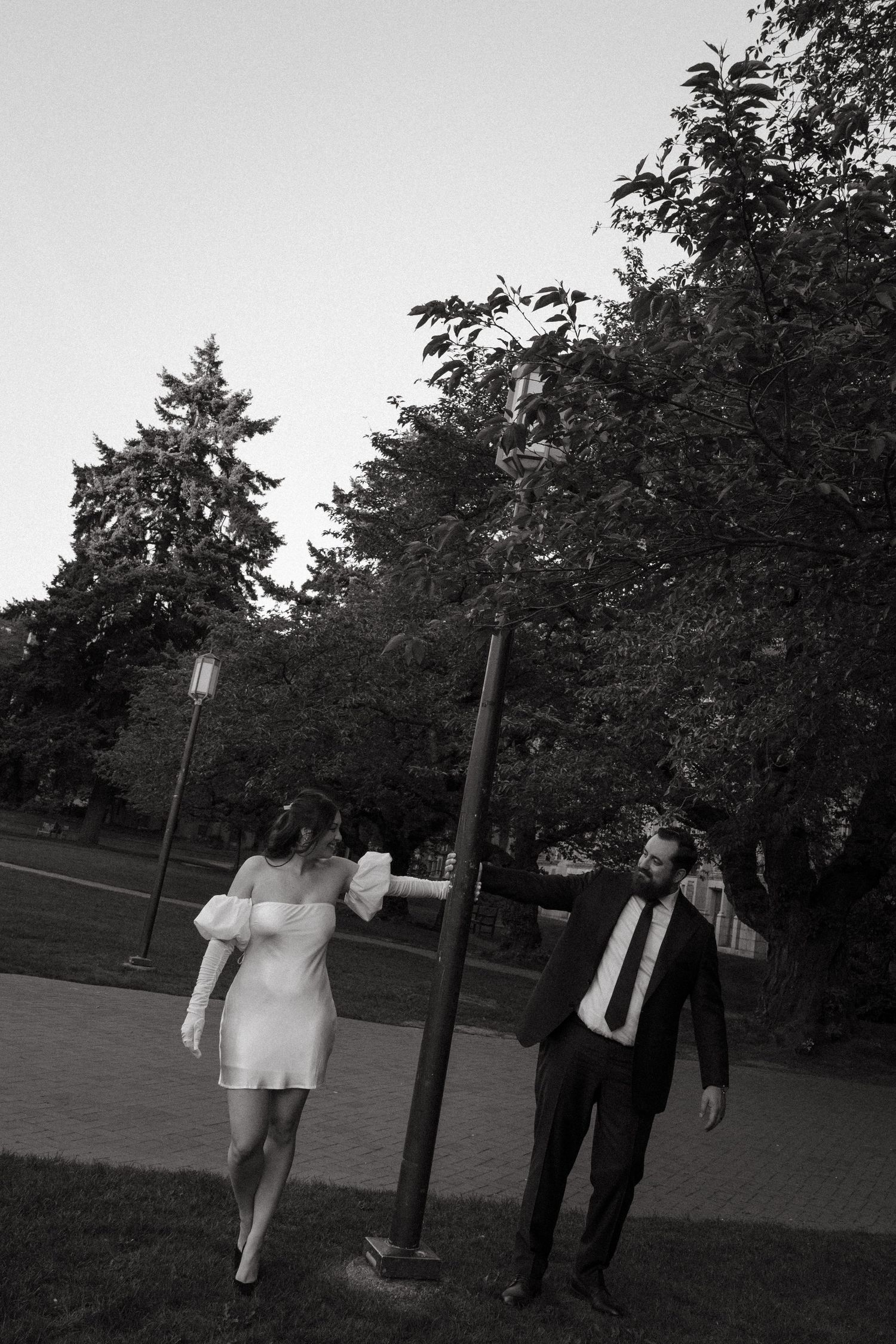 Black and white photo of a bride in white dress and groom in suit posing by a lamp post in a park.