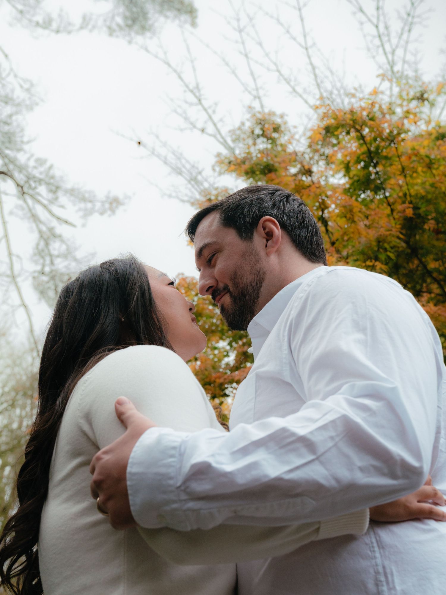 A smiling couple in white embraces and nearly kisses outdoors with colorful autumn foliage behind them.