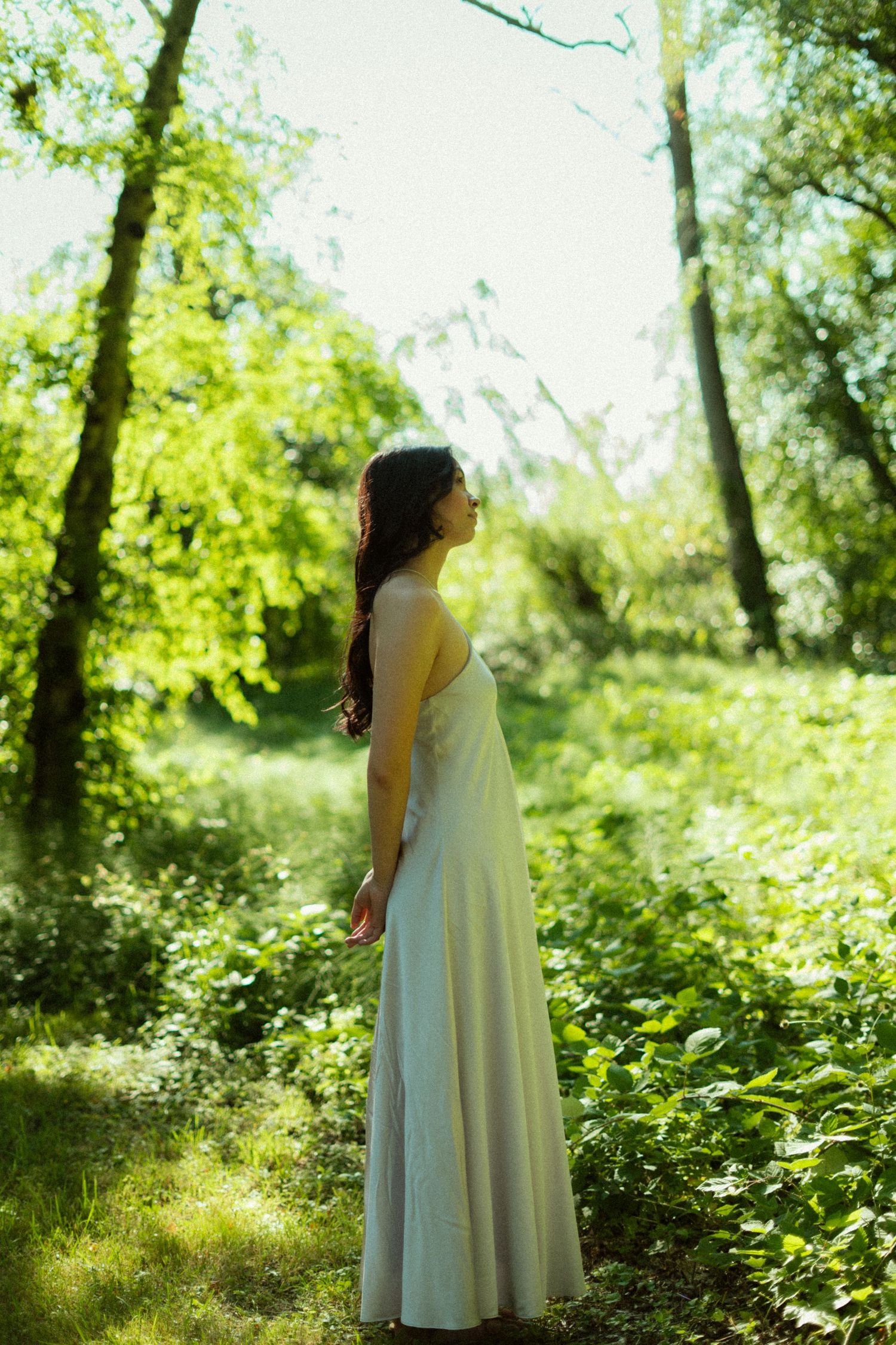 A woman in a long white dress stands peacefully in a lush green forest, bathed in soft sunlight filtering through the trees.