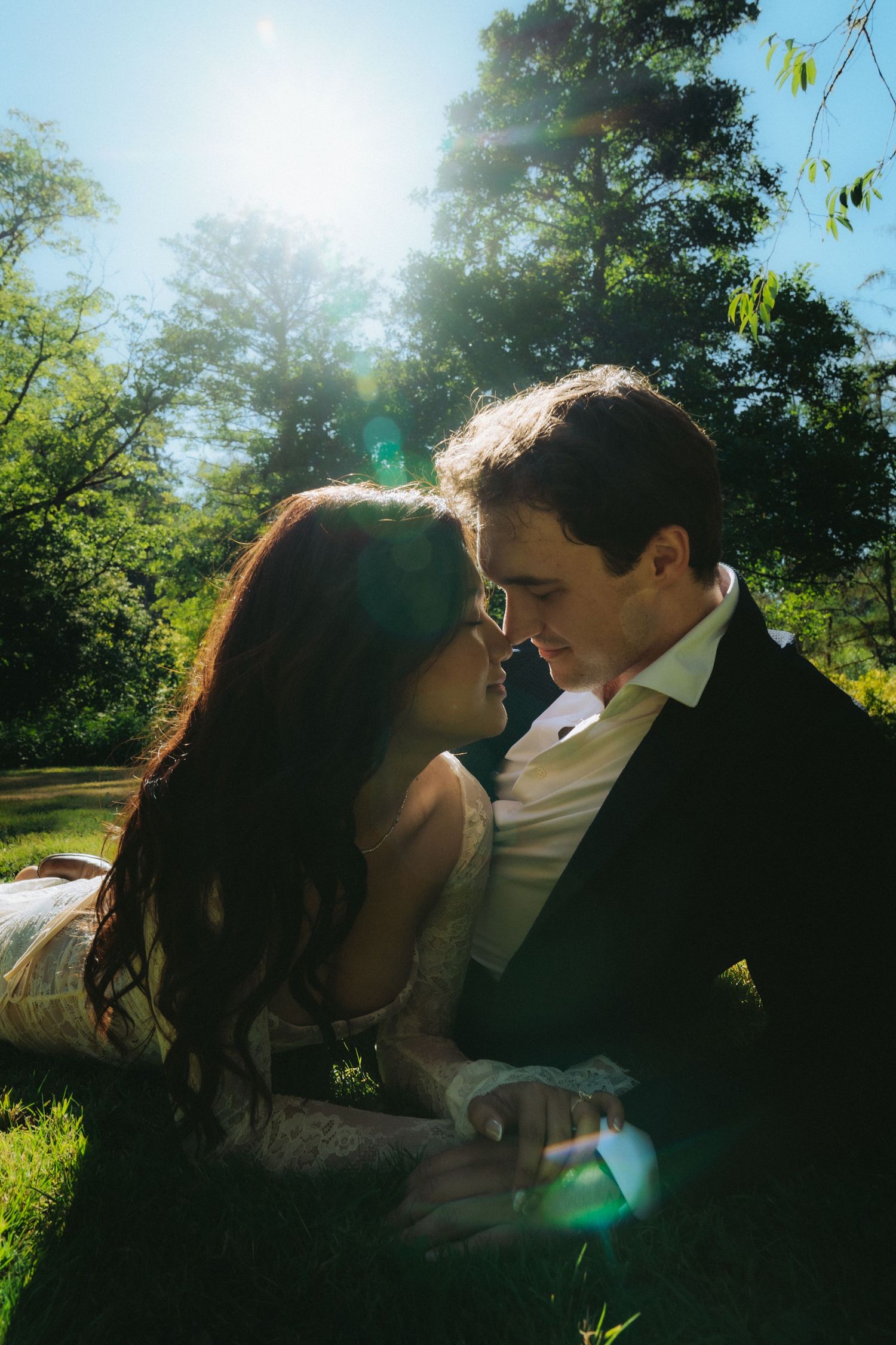 A couple in wedding attire shares a romantic moment lying on green grass, bathed in sunlight with lush trees behind them.