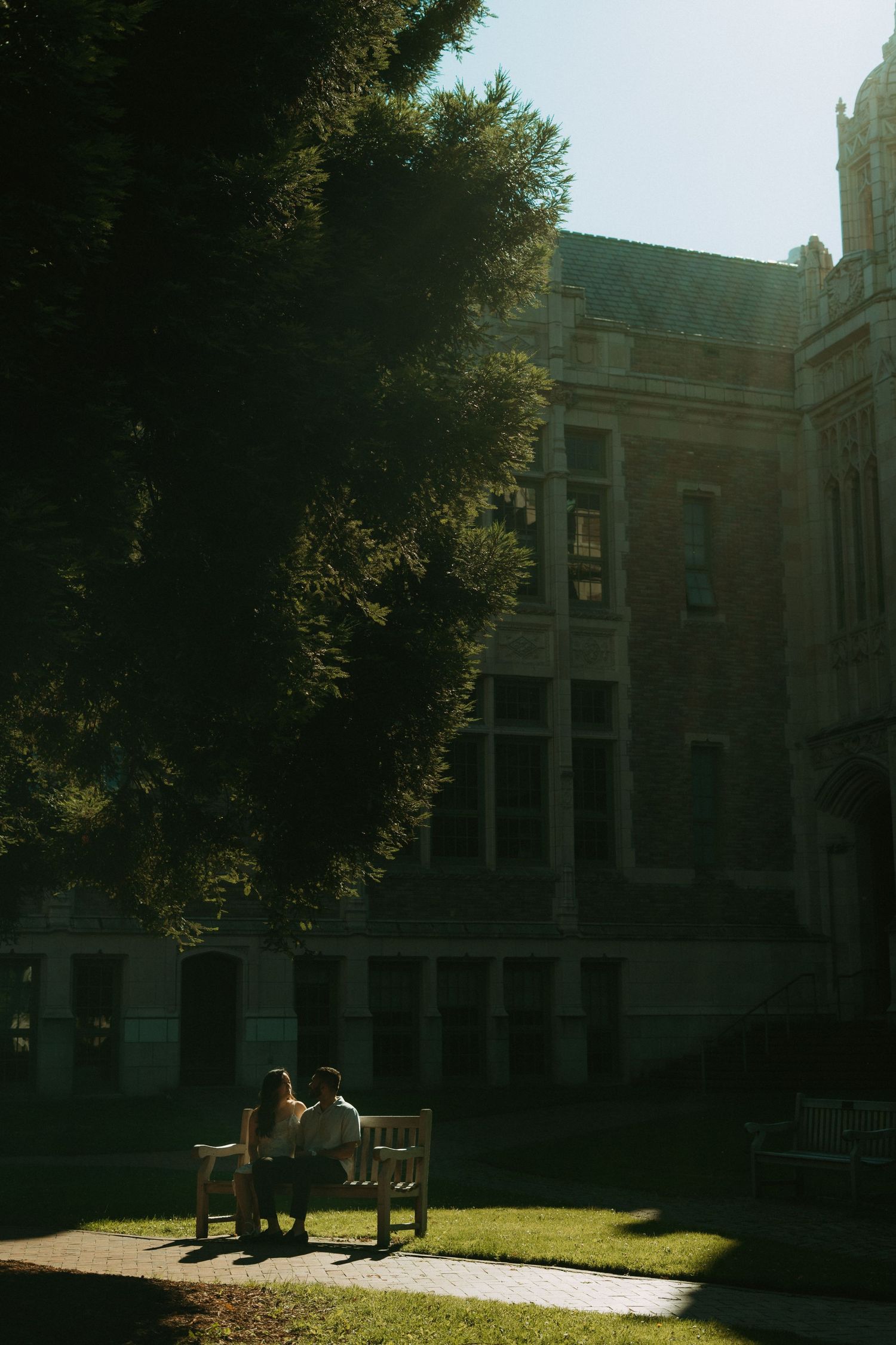 Couple sitting on a bench in sunlight outside a historic brick university building surrounded by lush green trees.