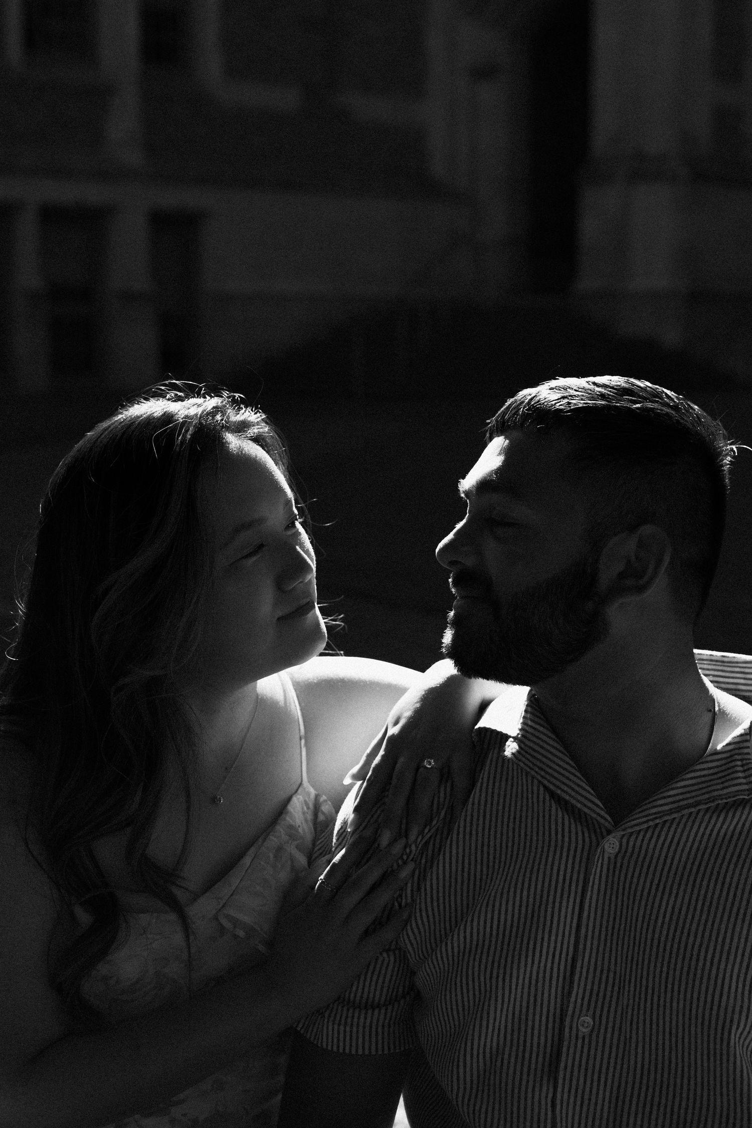 Black and white portrait of a bearded man and woman gazing at each other in dramatic side lighting.