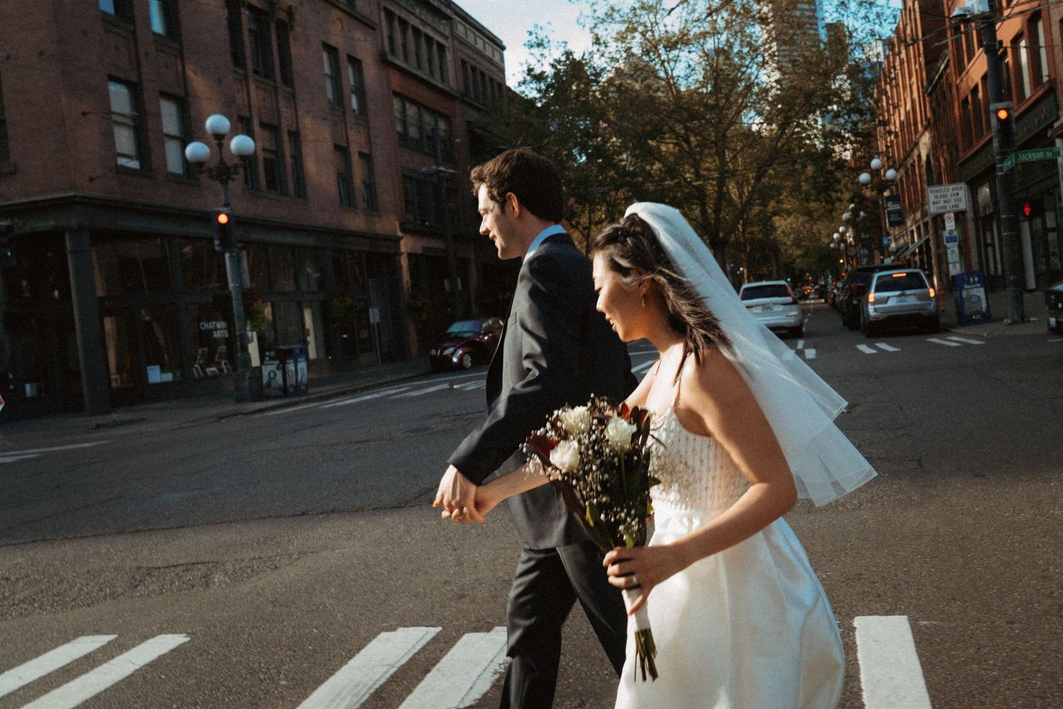 Bride and groom crossing a sunlit street in wedding attire, bride holding a bouquet, urban brick buildings in background.