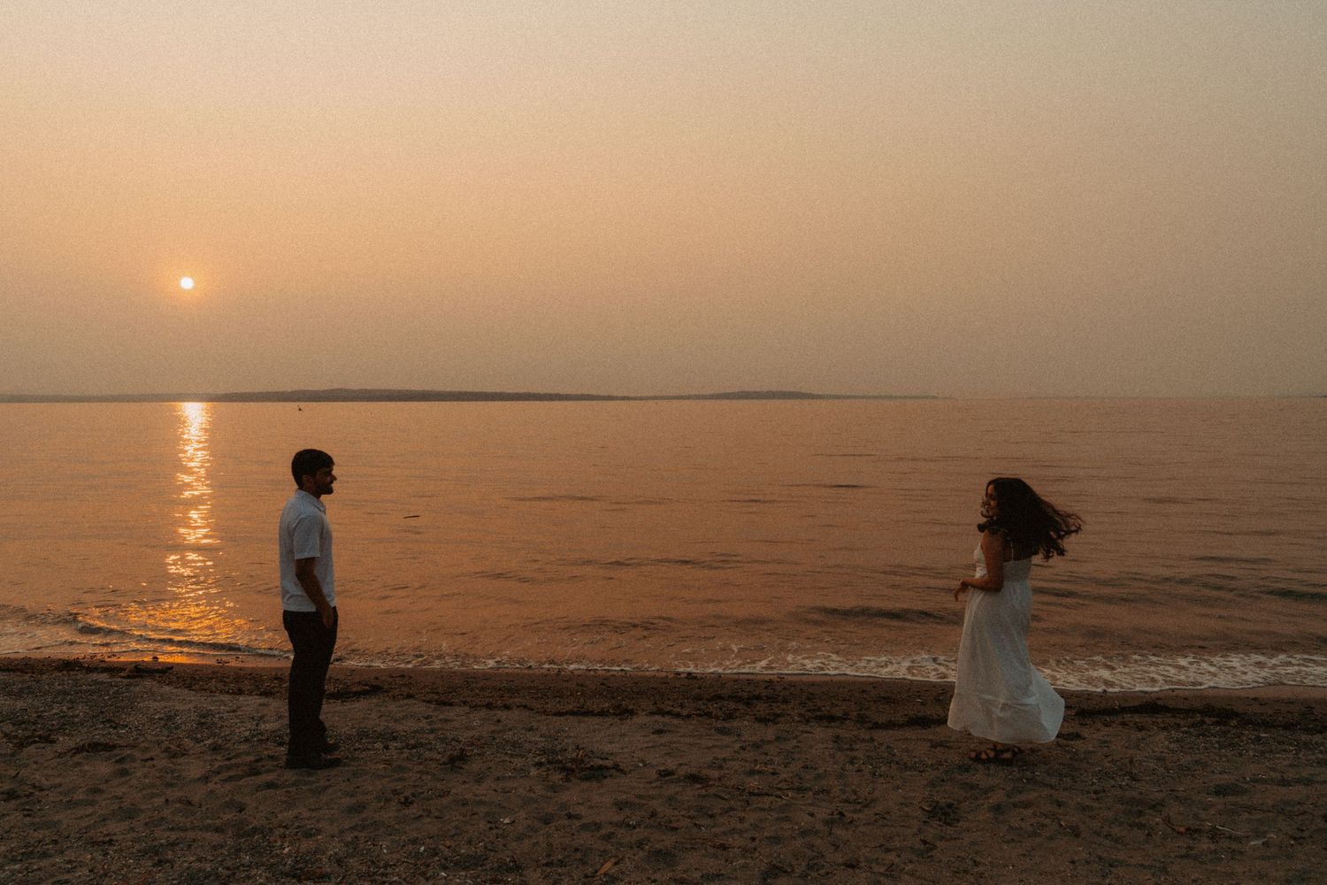 Couple standing apart on a sandy beach at sunset, with calm water and a warm orange sky in the background.