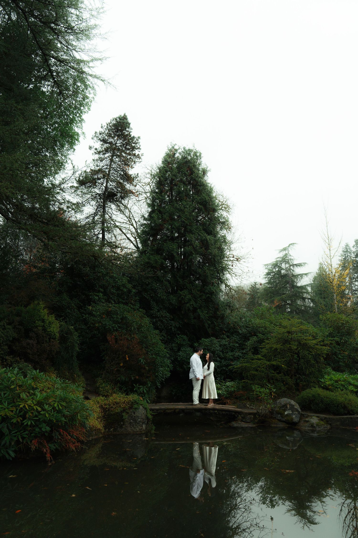 Couple sharing romantic moment on stone bridge reflected in tranquil pond in lush garden.