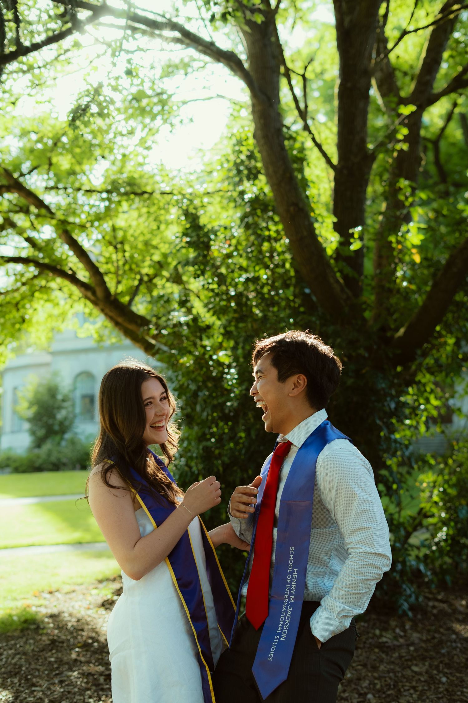 Two graduates in blue sashes smile at each other outdoors under green trees, celebrating their graduation day.
