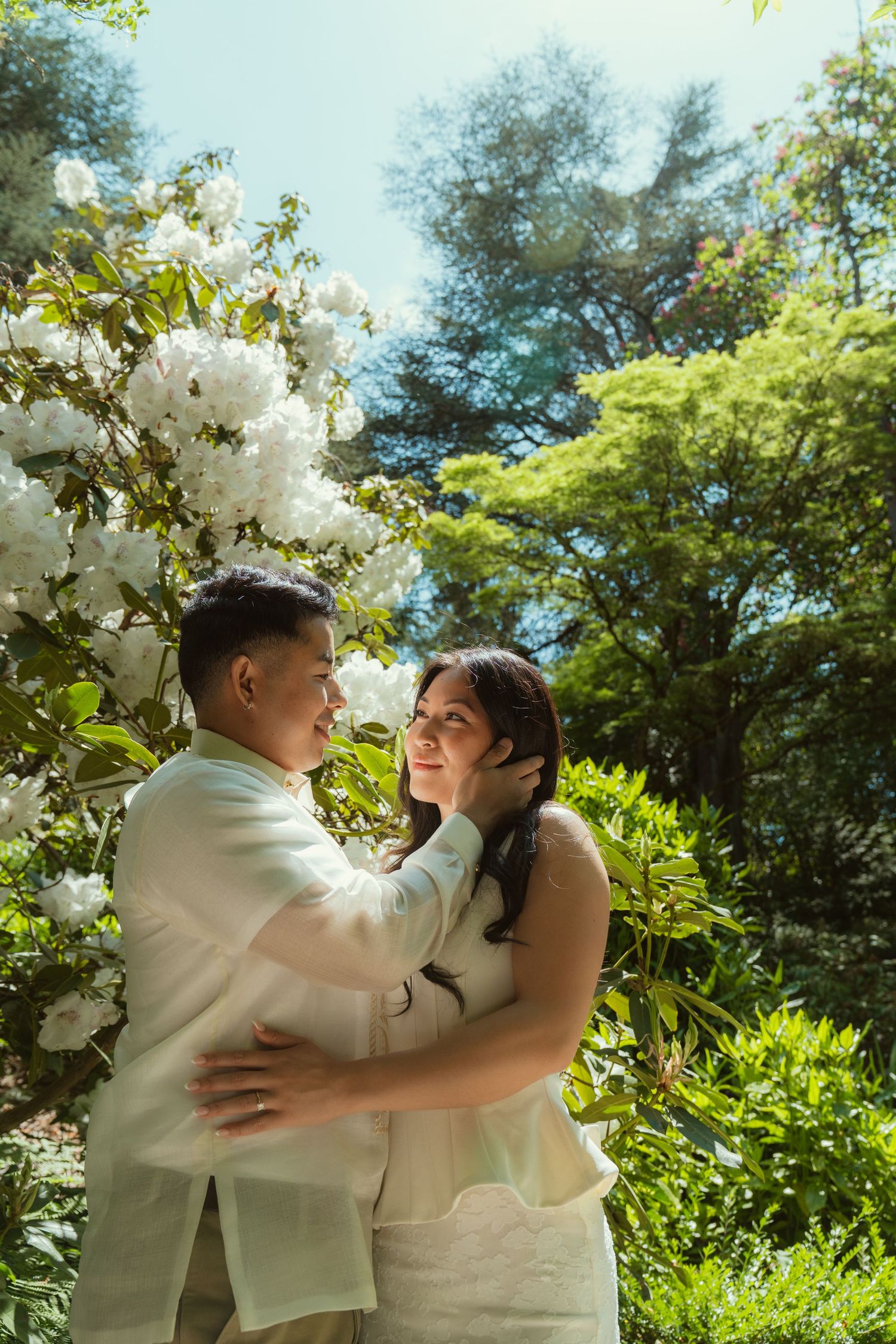 Couple embracing under white flowering tree in lush garden, wearing elegant cream outfits on sunny day.