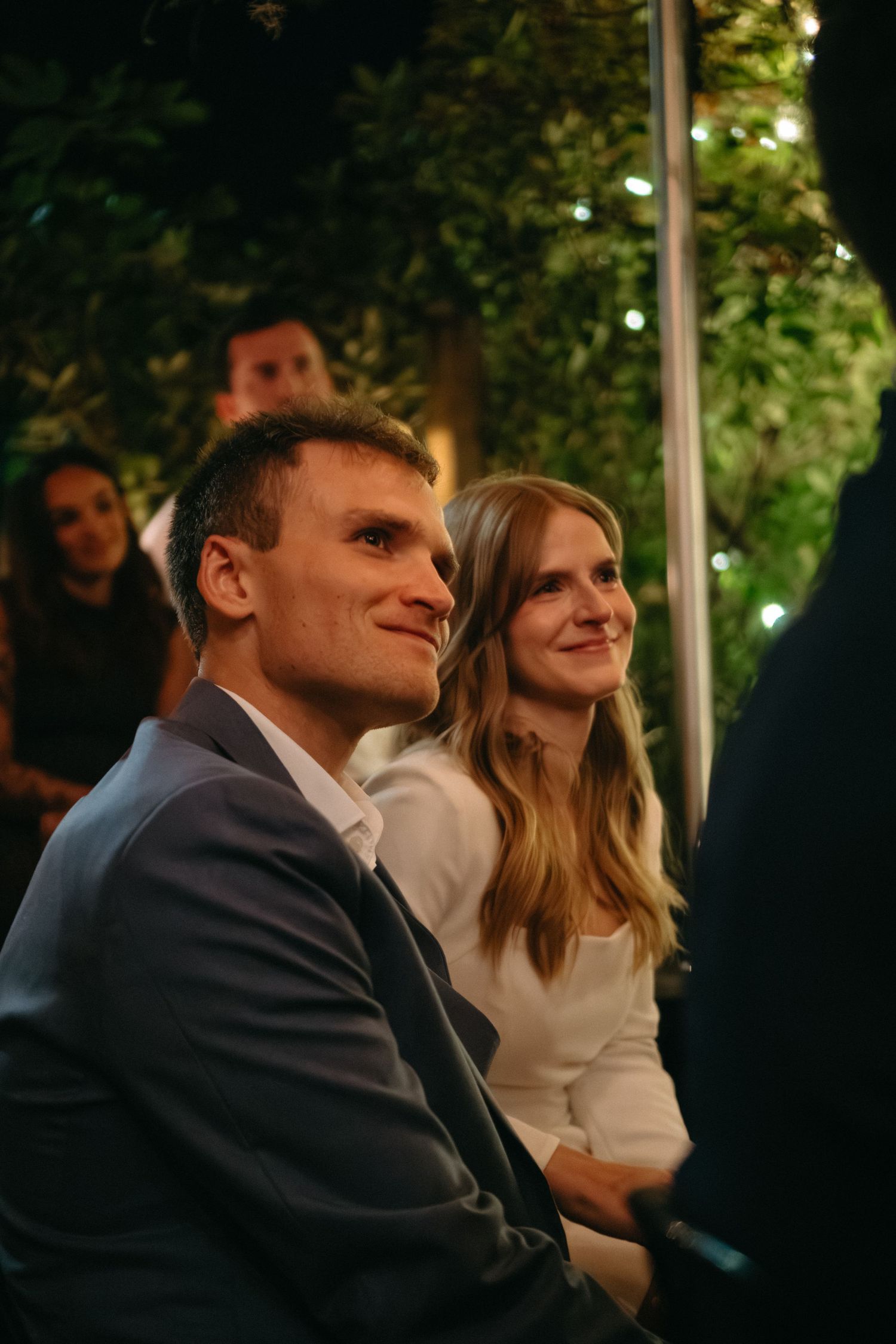 Happy couple smiling at an outdoor garden wedding ceremony with twinkling lights and greenery.