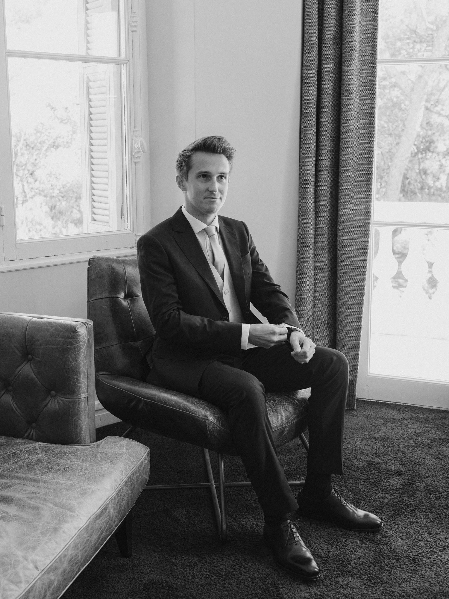 Man in dark suit sitting in leather chair by window, black and white portrait photography.