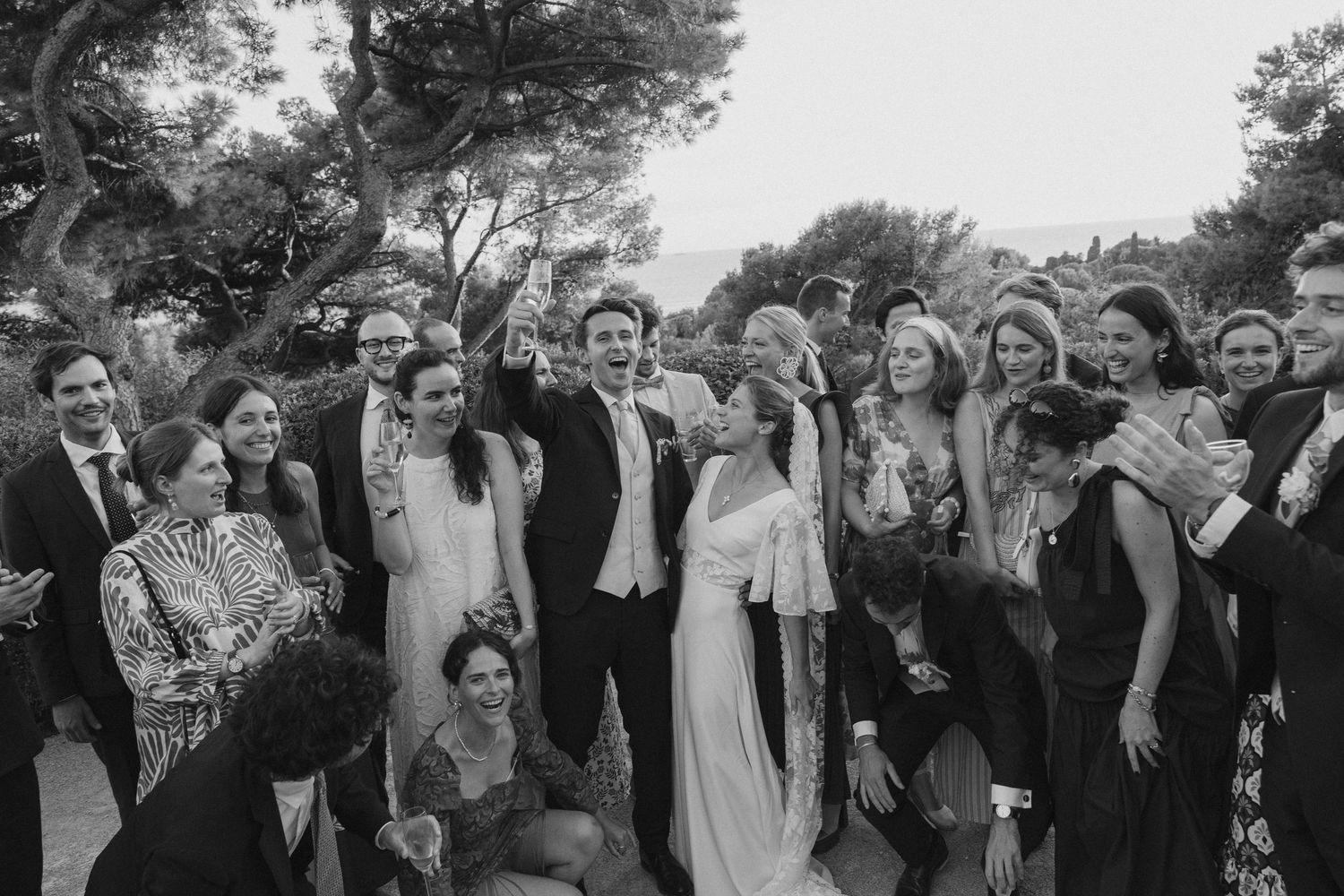 Black and white wedding group photo with bride and groom celebrating joyfully outdoors with guests.