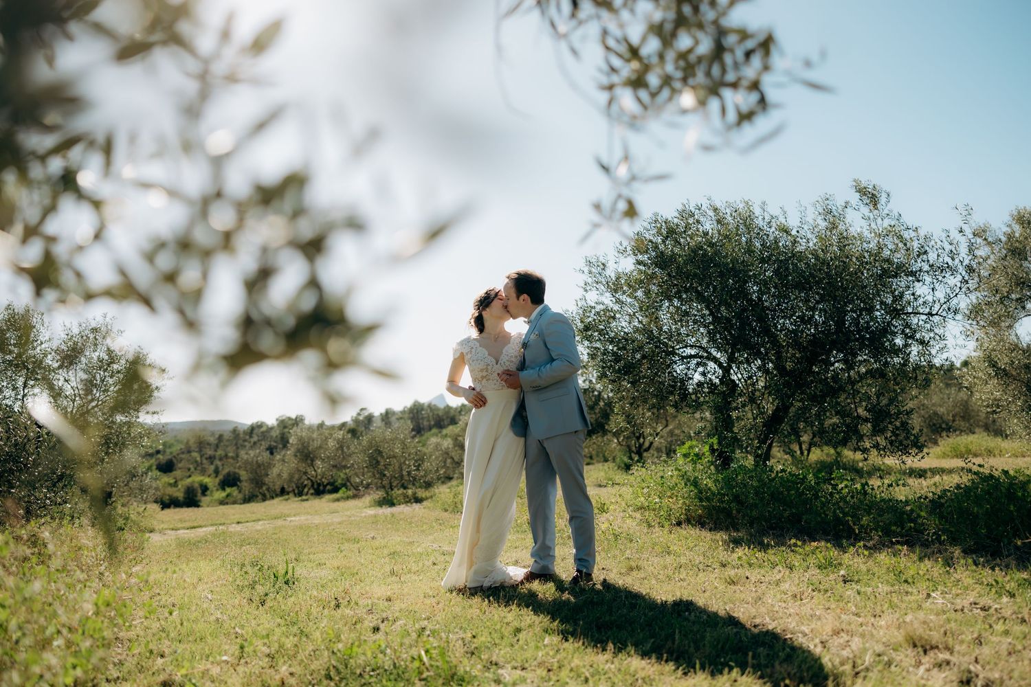 A couple embraces on a sunny hillside surrounded by lush green foliage and natural scenery.