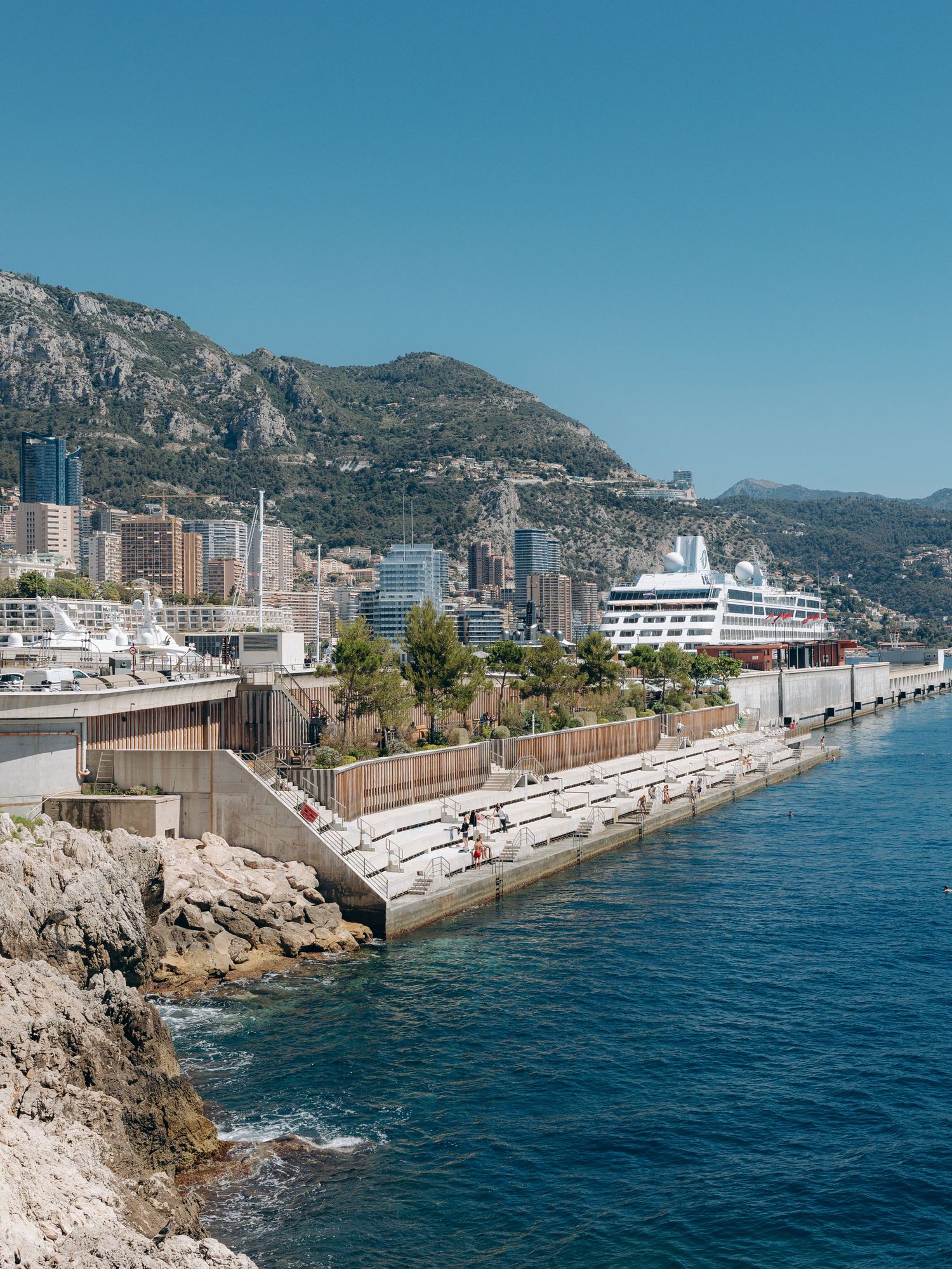 Scenic Monaco coastline with cruise ship, luxury buildings, and lush mountains under a clear blue sky.