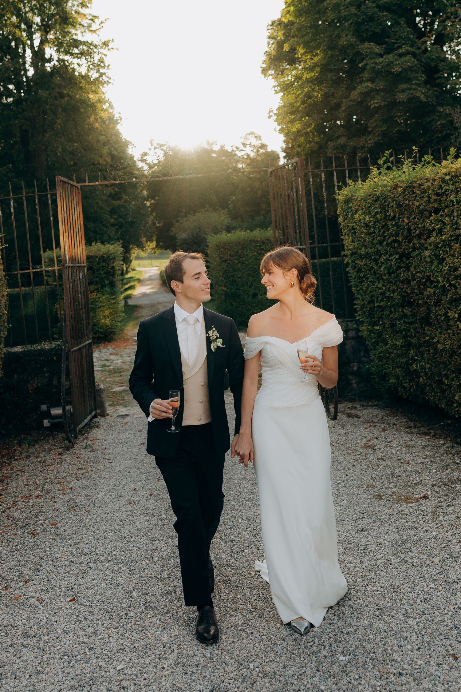 A couple in wedding attire walks together on a gravel path at sunset while sharing a romantic moment.