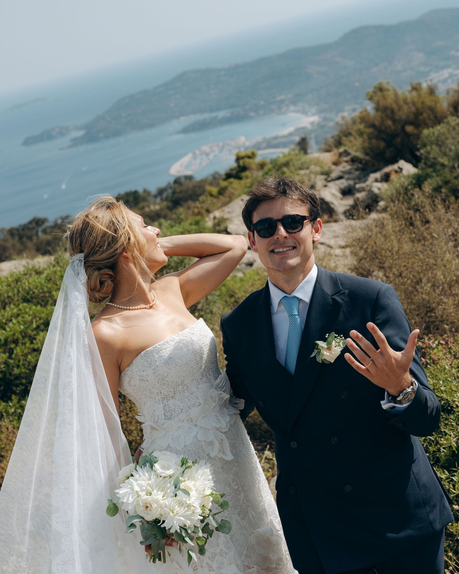 Bride and groom posing on a scenic coastal hilltop, she holds white bouquet while he waves showing wedding ring.