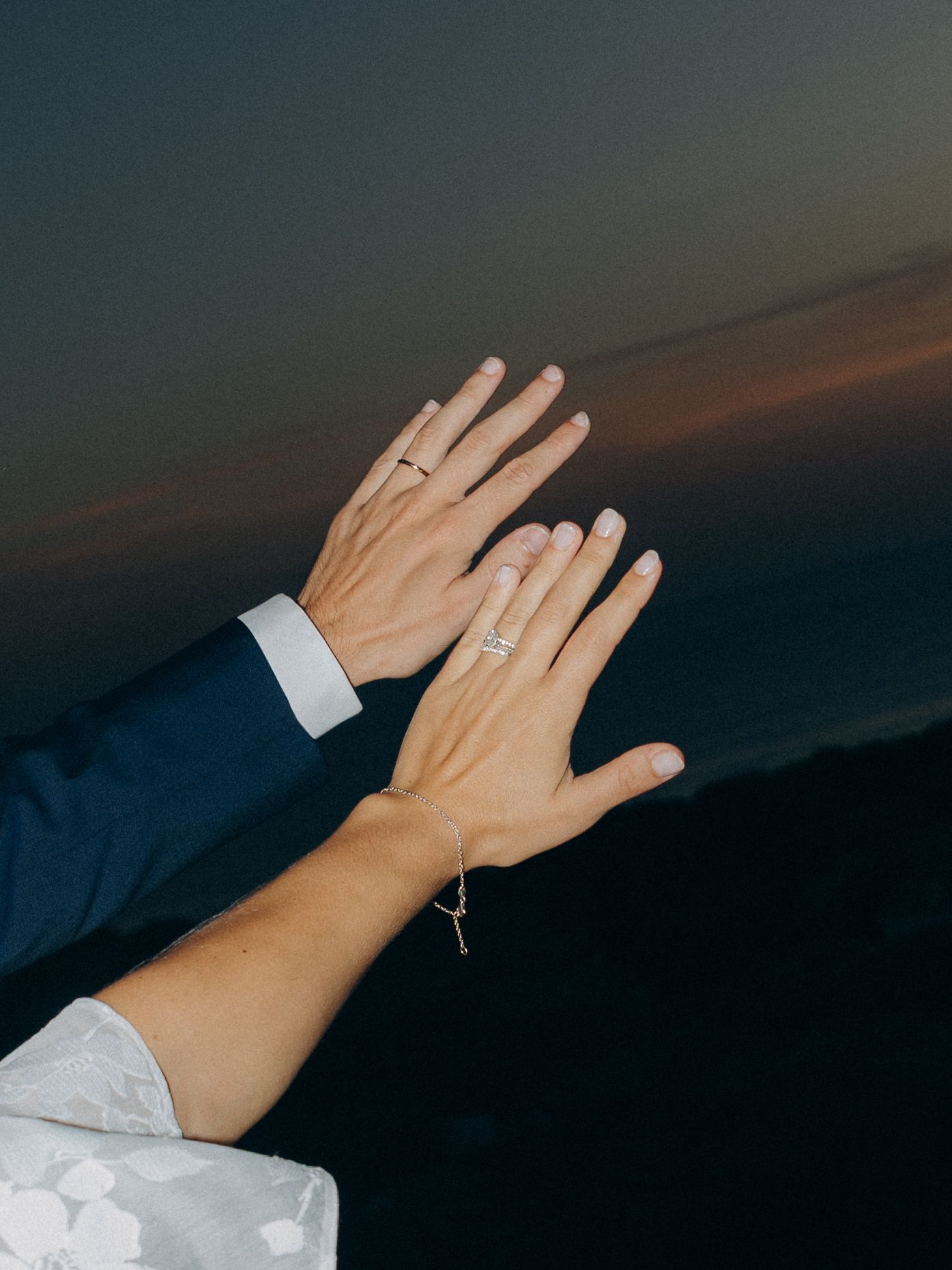 Couple raising hands at sunset showing wedding rings, bride in white dress and groom in navy suit.