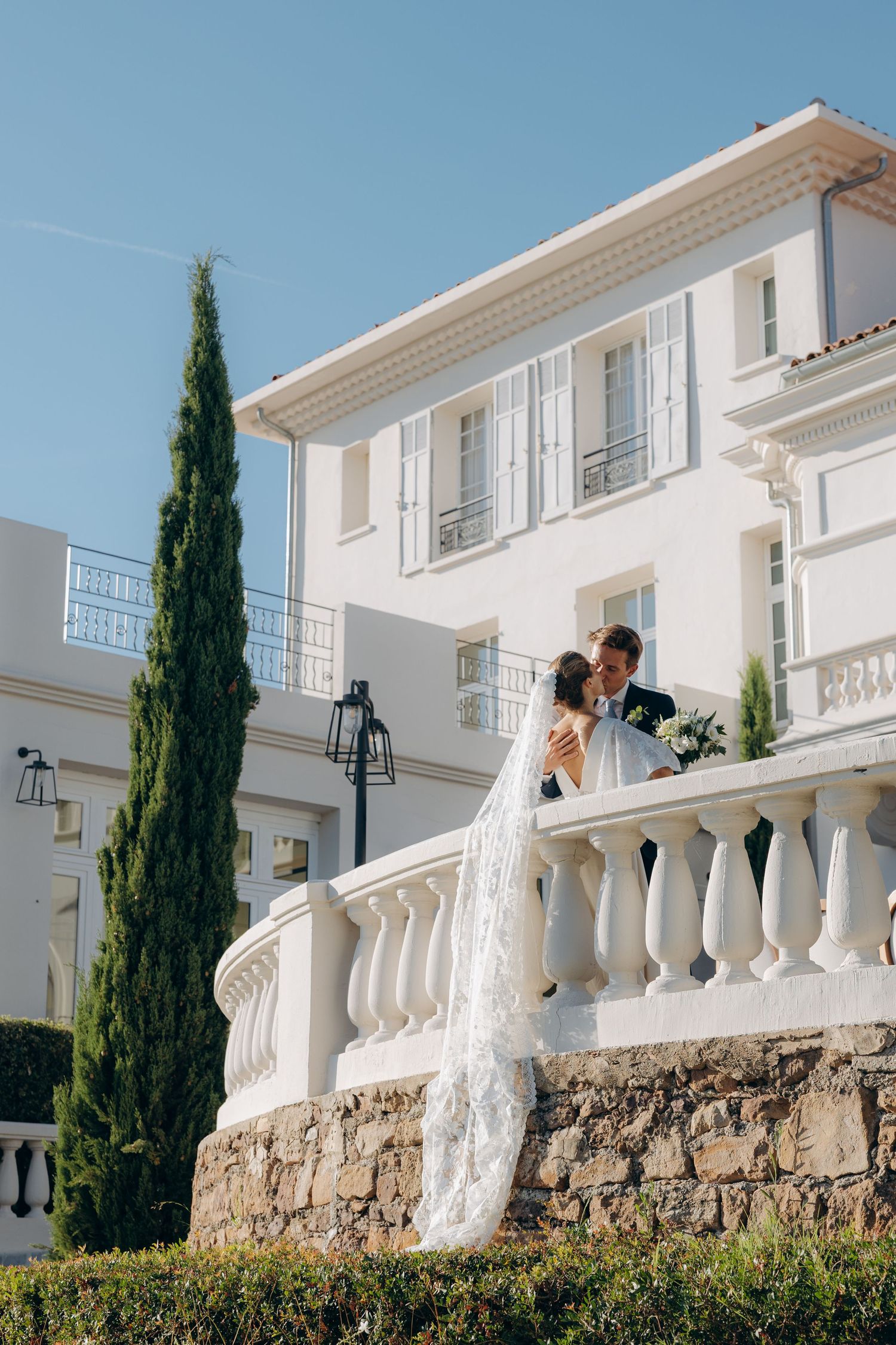 Bride and groom share a romantic kiss on a white balustrade terrace of an elegant white villa under a clear blue sky.