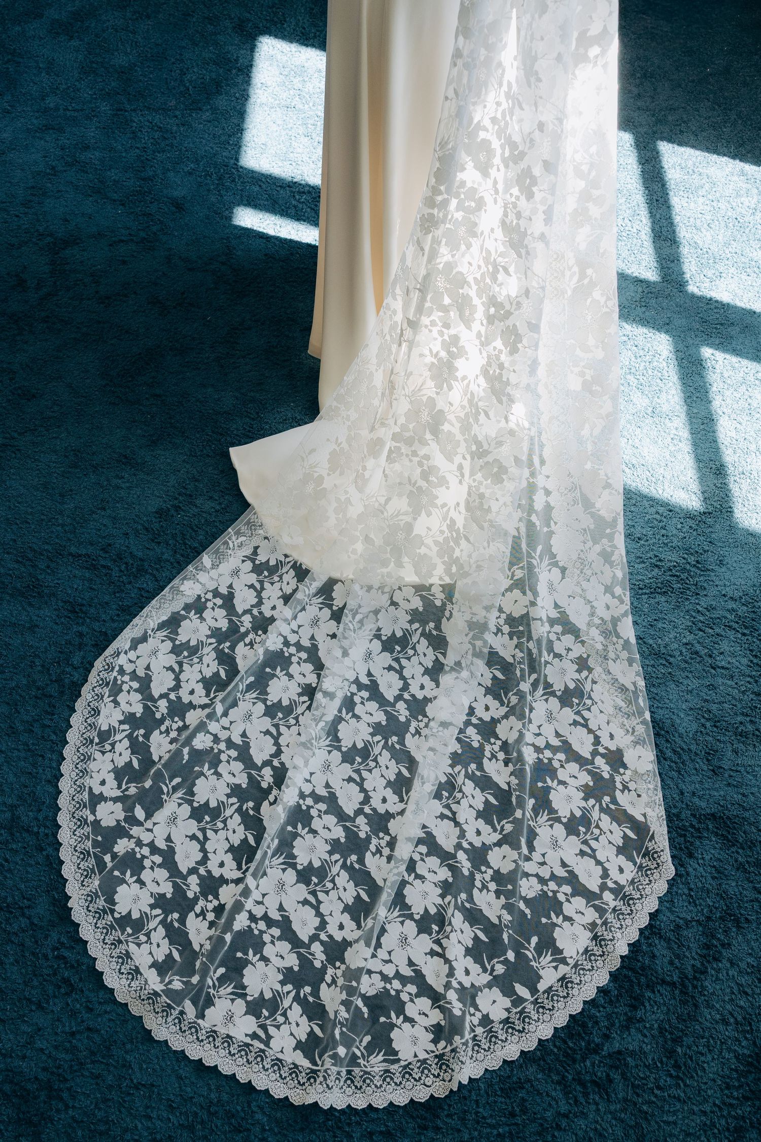 Elegant white lace bridal veil with floral pattern trailing on teal carpet in soft window light.