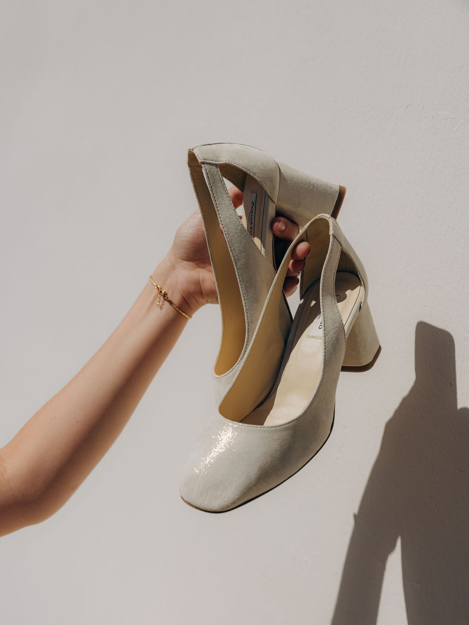 A hand holding elegant gold metallic block heel pumps against a white wall in natural light.