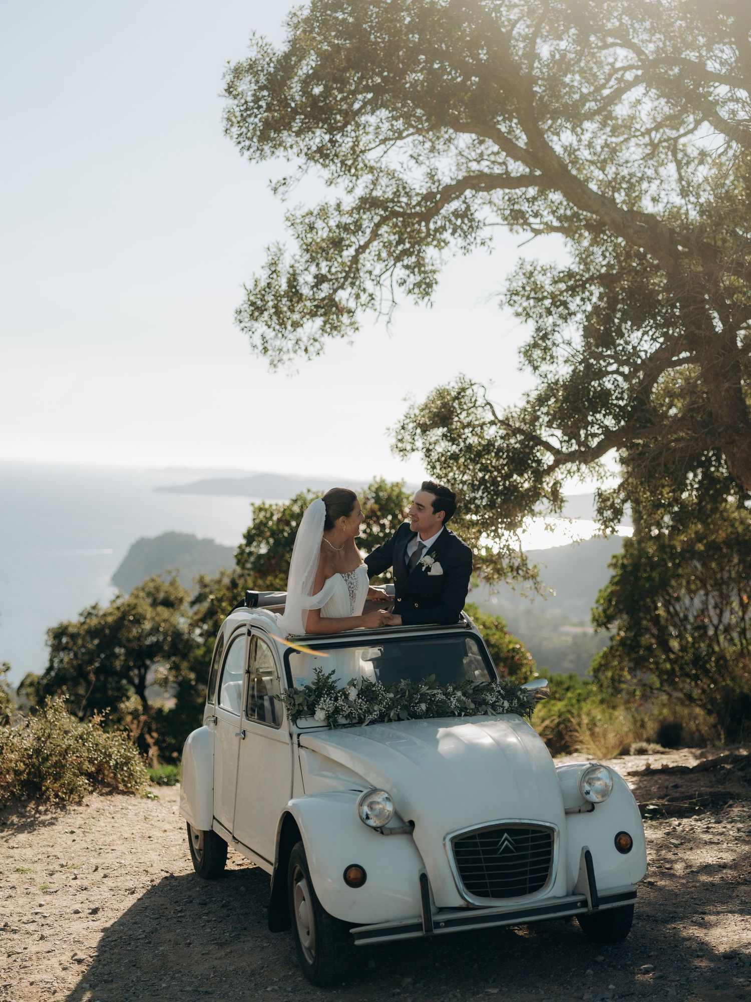 Vintage white Citroen car parked under trees with mountains in the background.