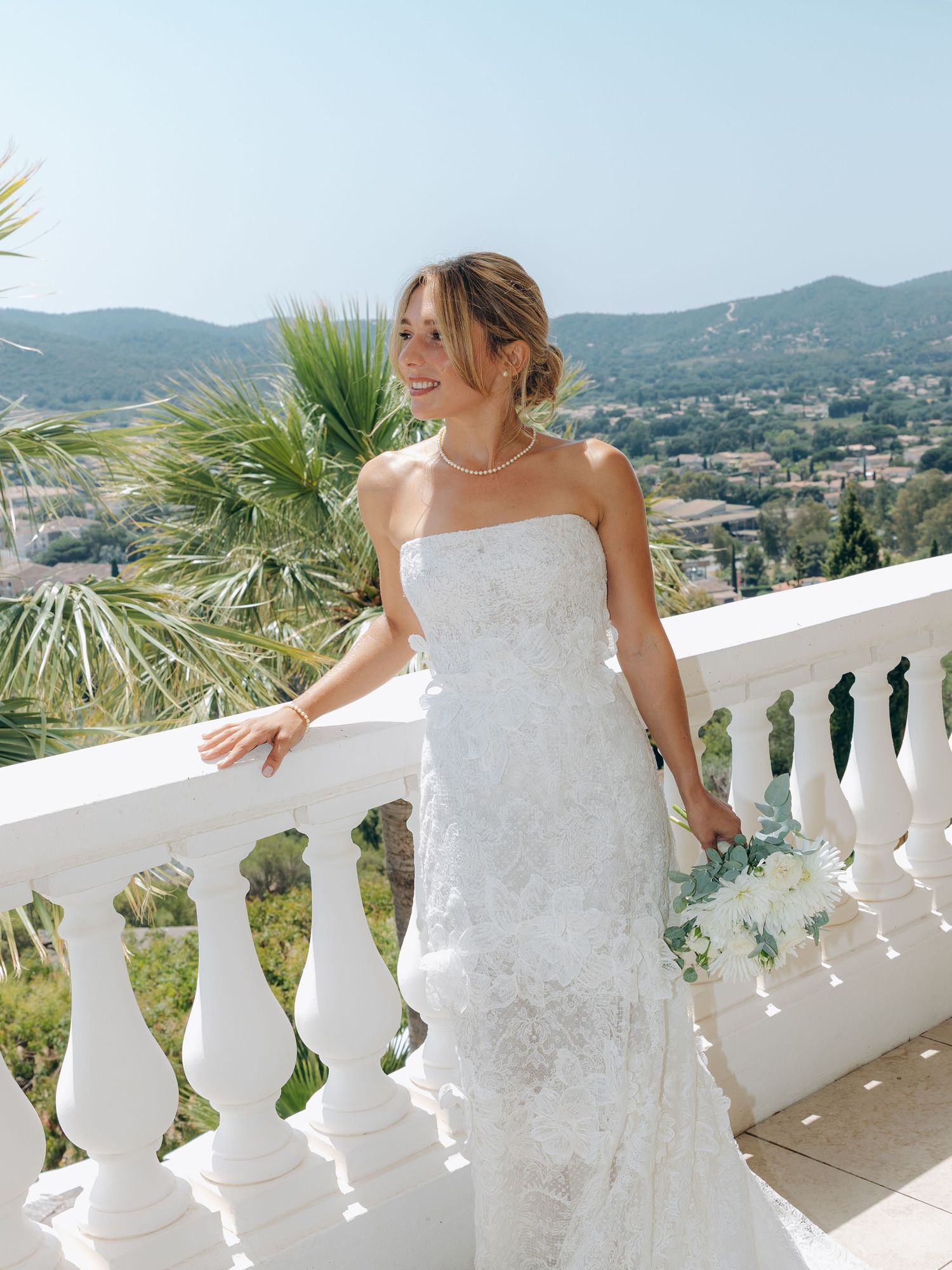 Bride in strapless lace wedding gown holding bouquet on white balcony with mountain views.