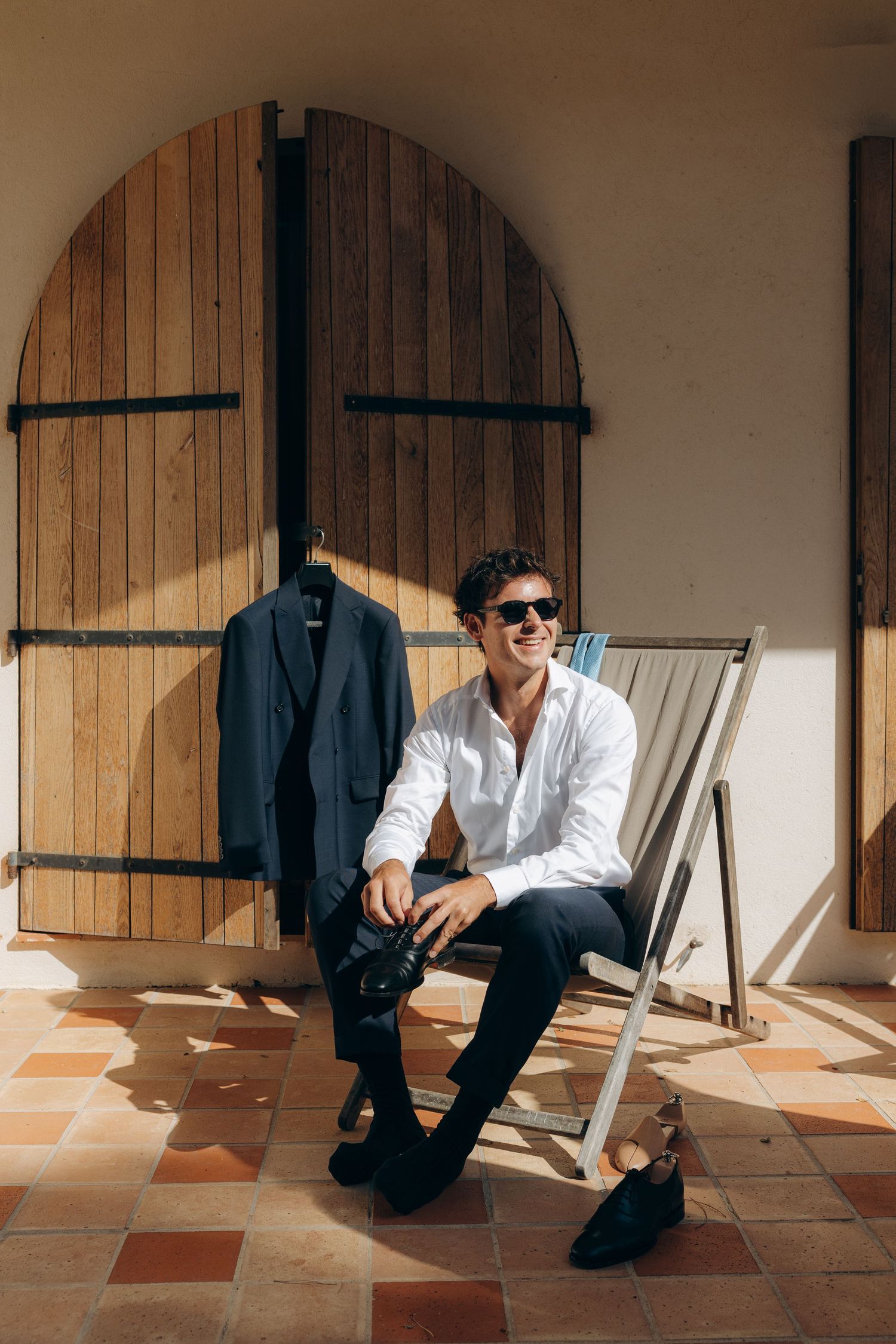 Smiling man in white shirt and sunglasses sits in deck chair outdoors, suit jacket hanging on wooden arched door behind him.