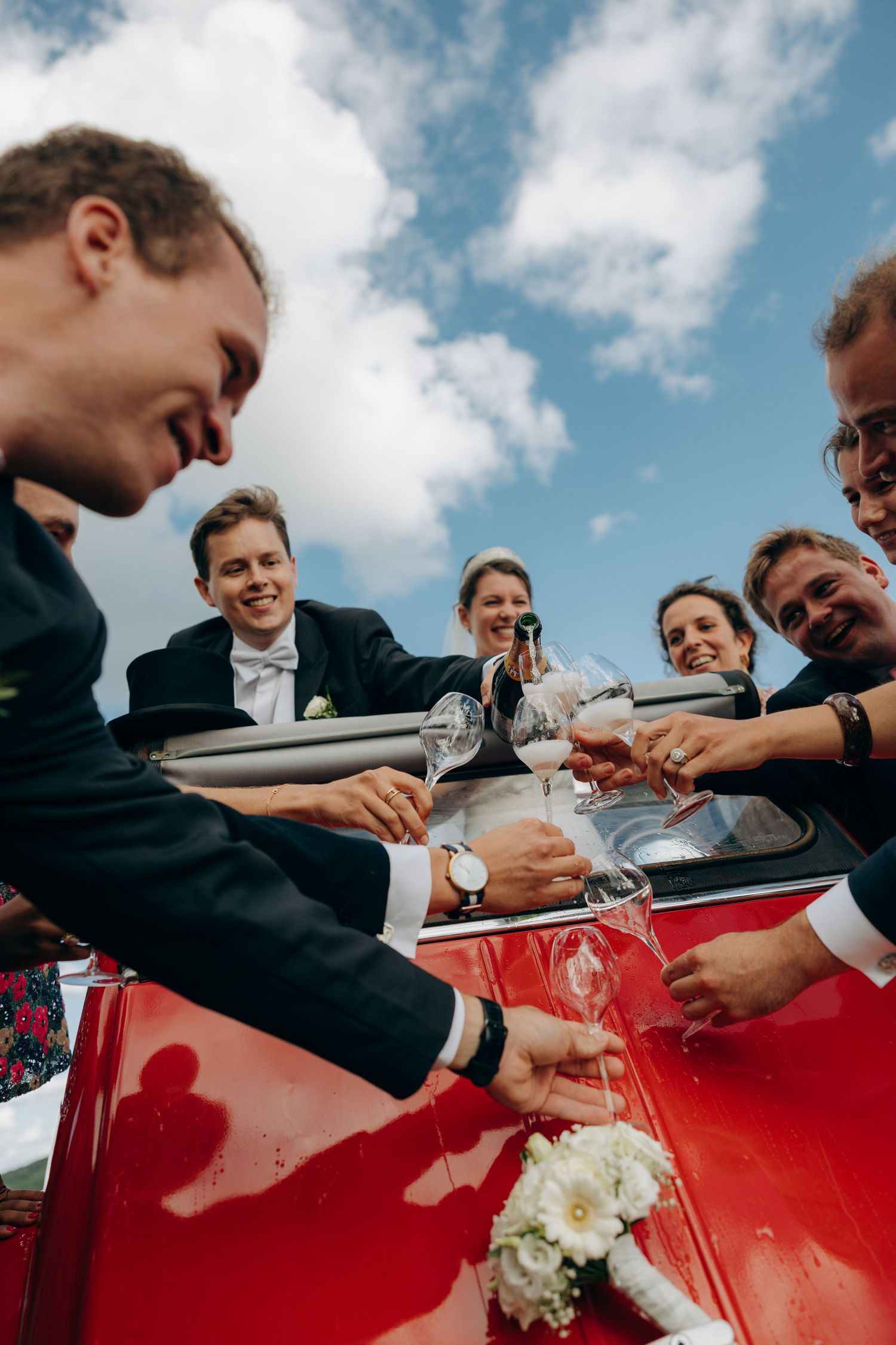 A group in formal attire signs a red classic car at an outdoor celebration under blue skies with white clouds.