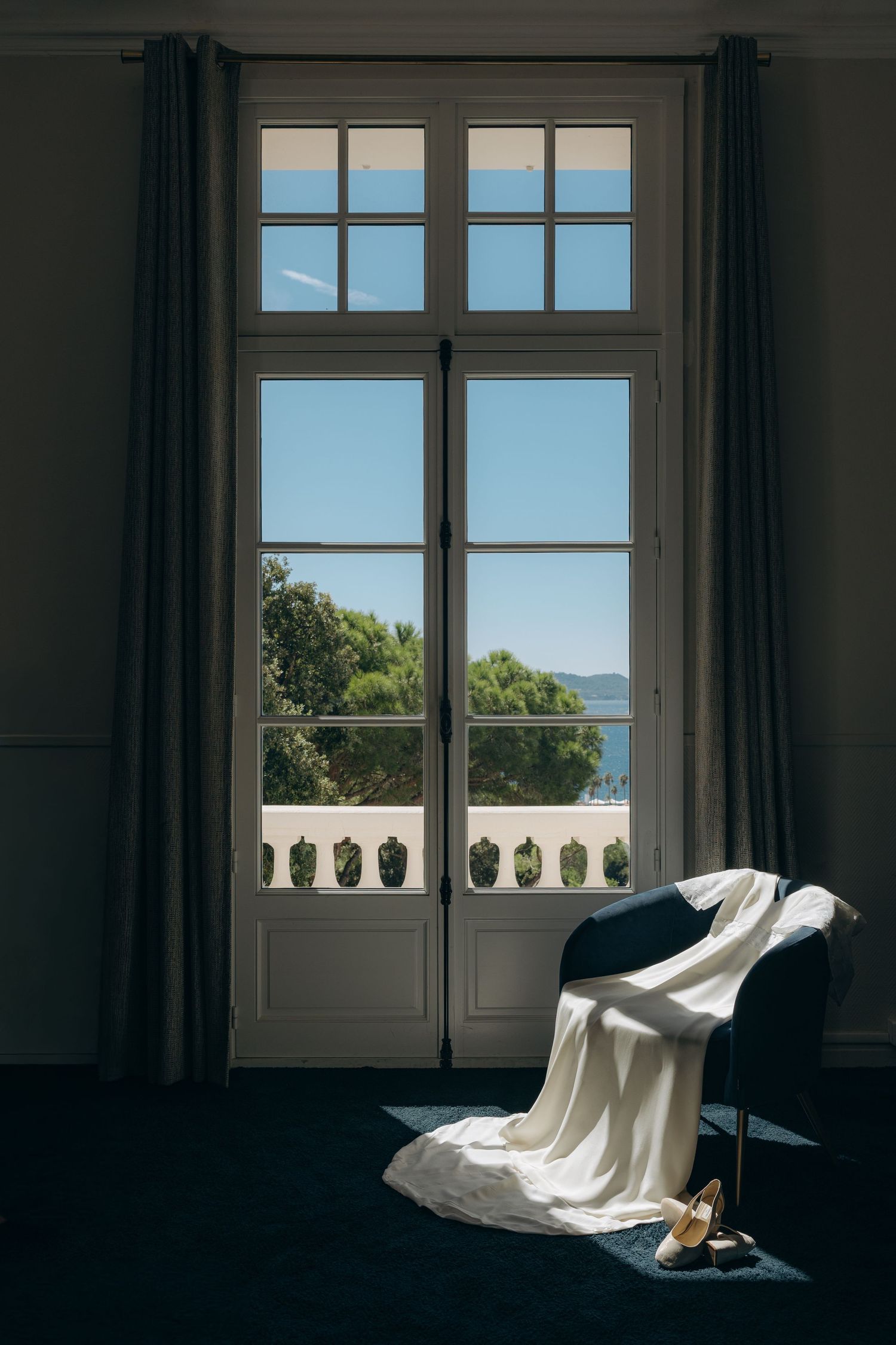 Elegant French doors open to a balcony with sea views, a white draped chair and heels in soft natural light.