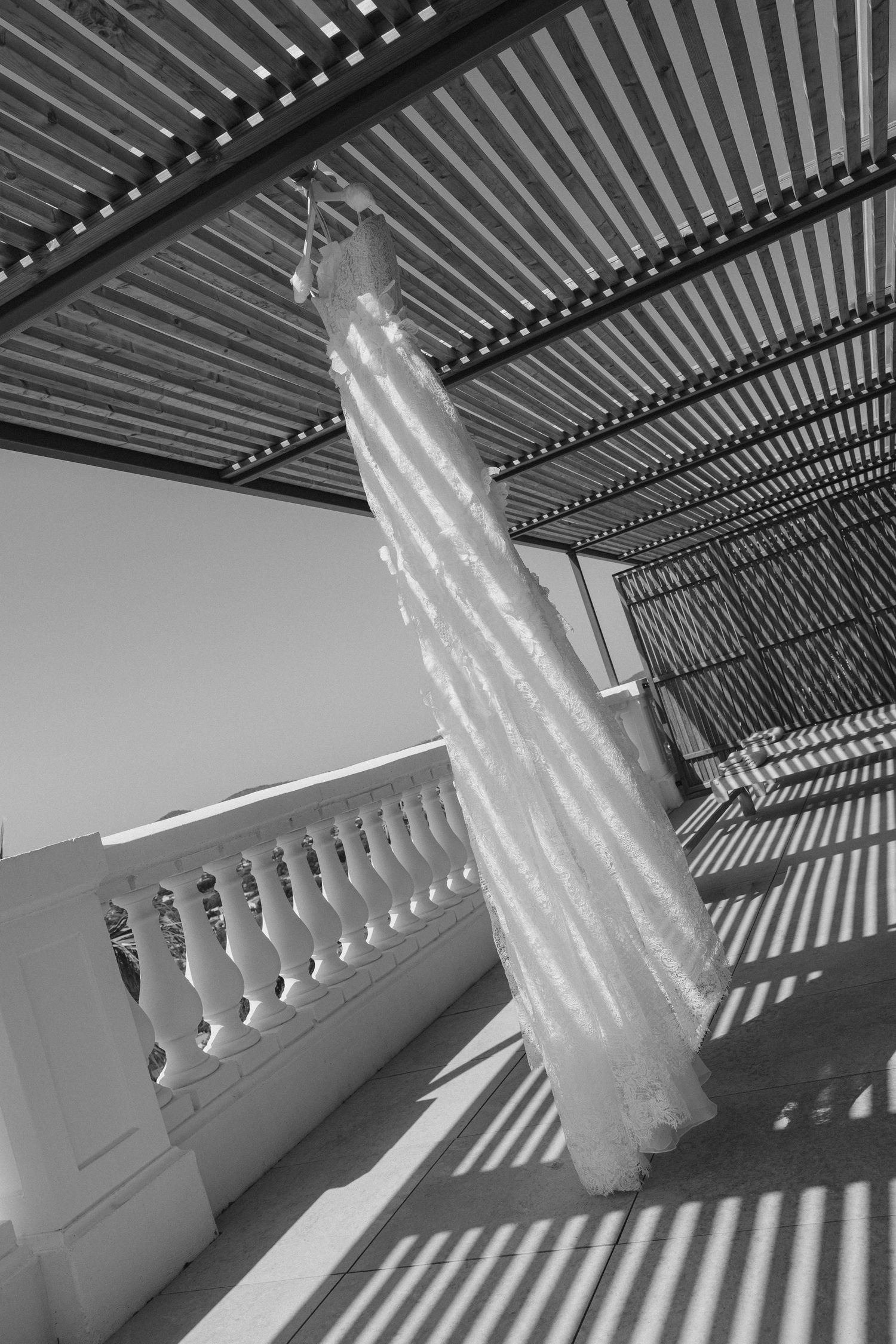 Black and white photo of an elegant wedding dress hanging under a pergola on a sunlit balcony with decorative railings.