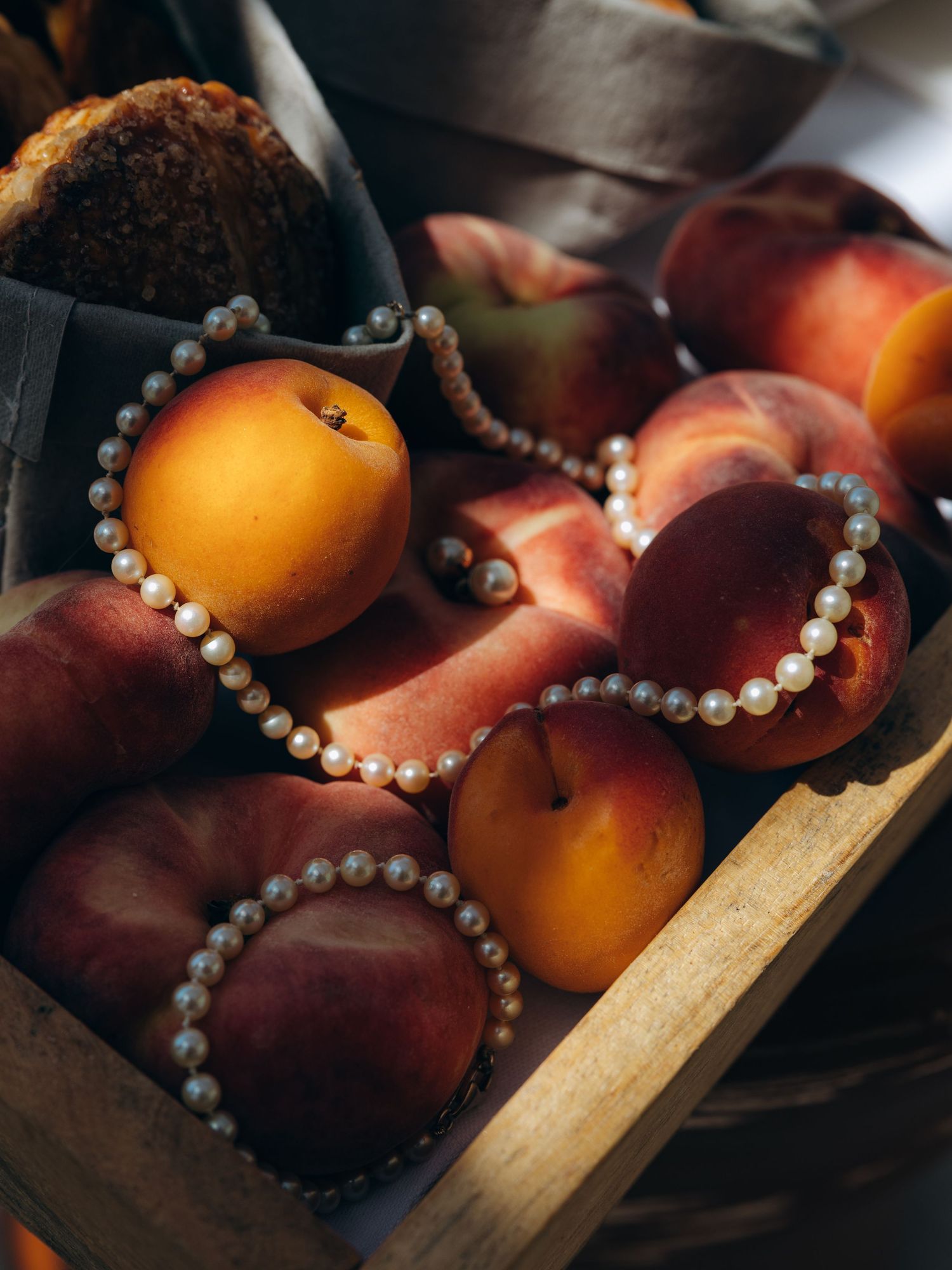 A rustic wooden crate filled with ripe peaches adorned with elegant pearl necklaces in warm natural light.