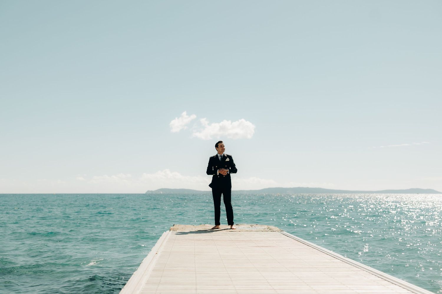 A scenic panorama of a Mediterranean coastline with turquoise waters and a wooden pier extending into the sea.