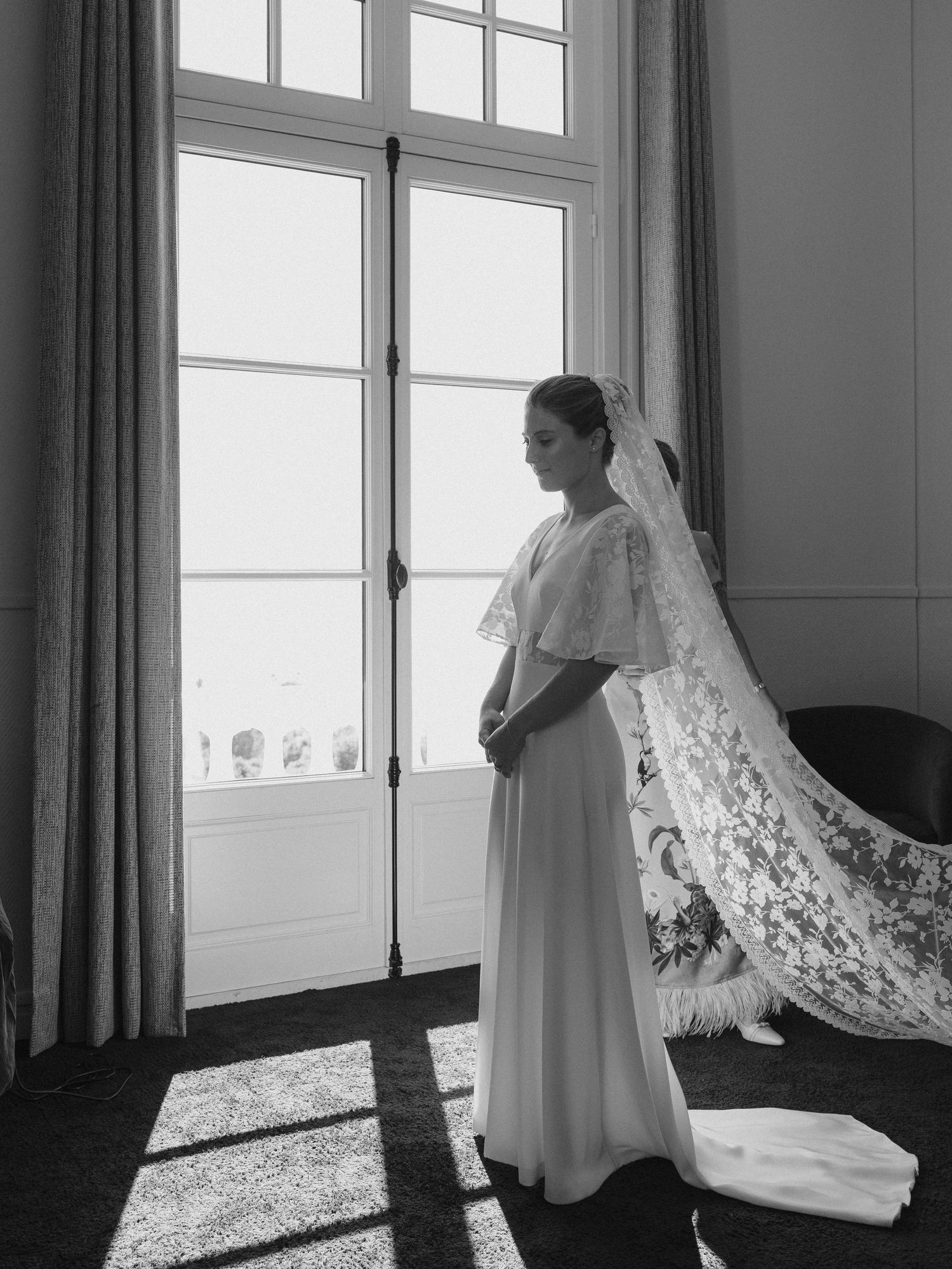 Black and white photo of a bride in a flowing gown and lace veil standing by tall French doors in soft natural light.