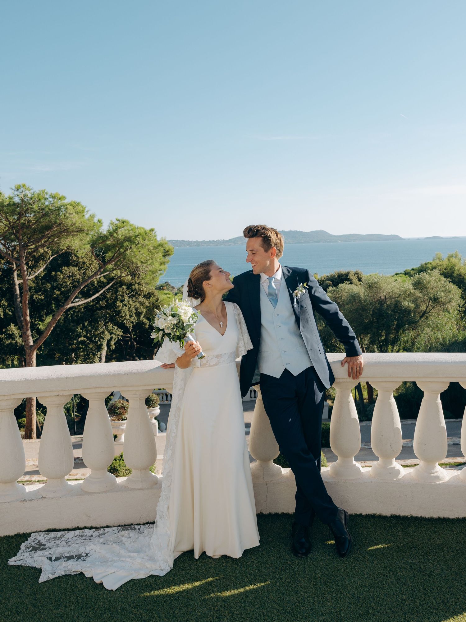 Bride and groom share a romantic moment on a white balcony terrace with scenic mountain and sea views.