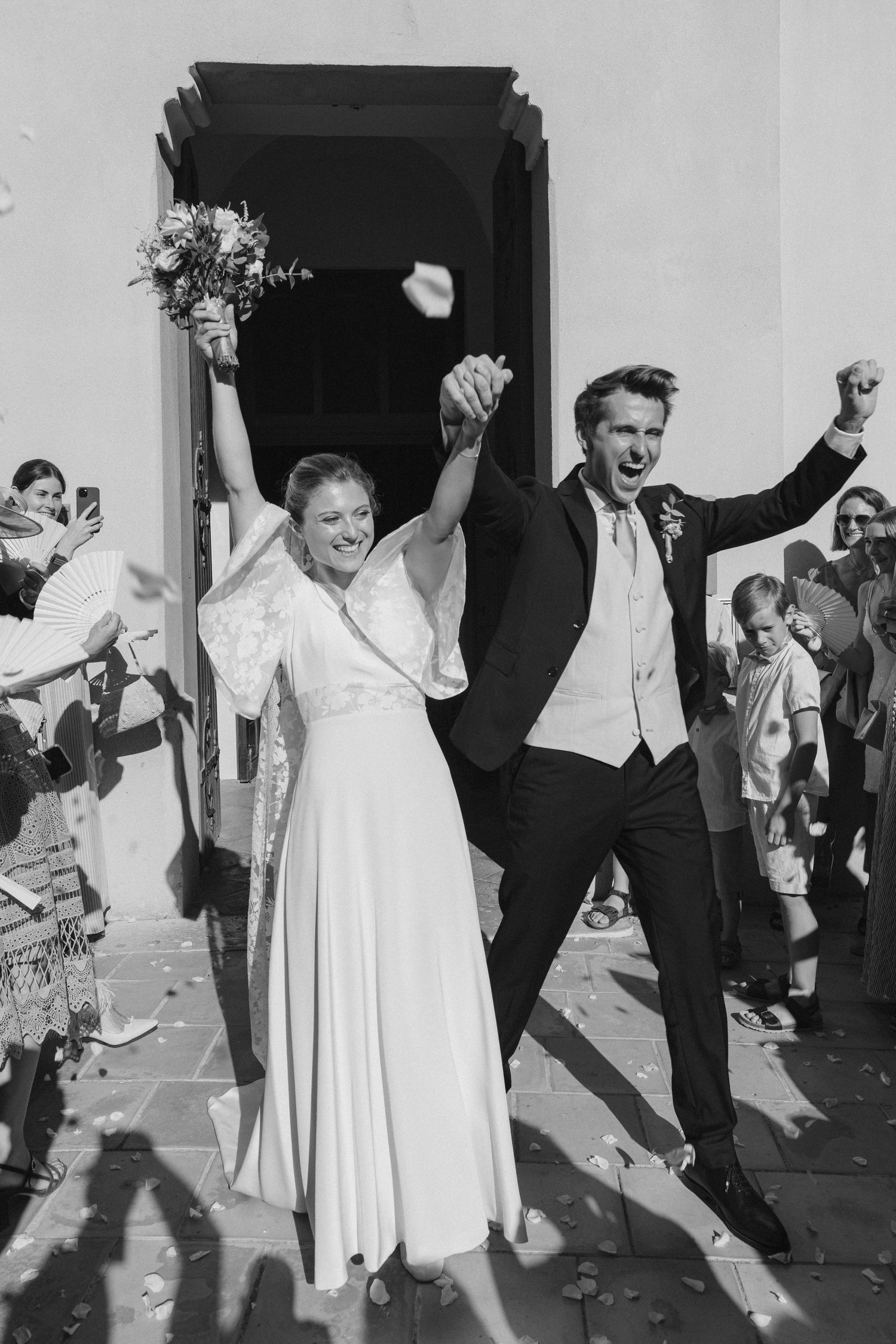 Black and white photo of joyful newlyweds celebrating outside church, bride holding bouquet, groom jumping with fist raised.